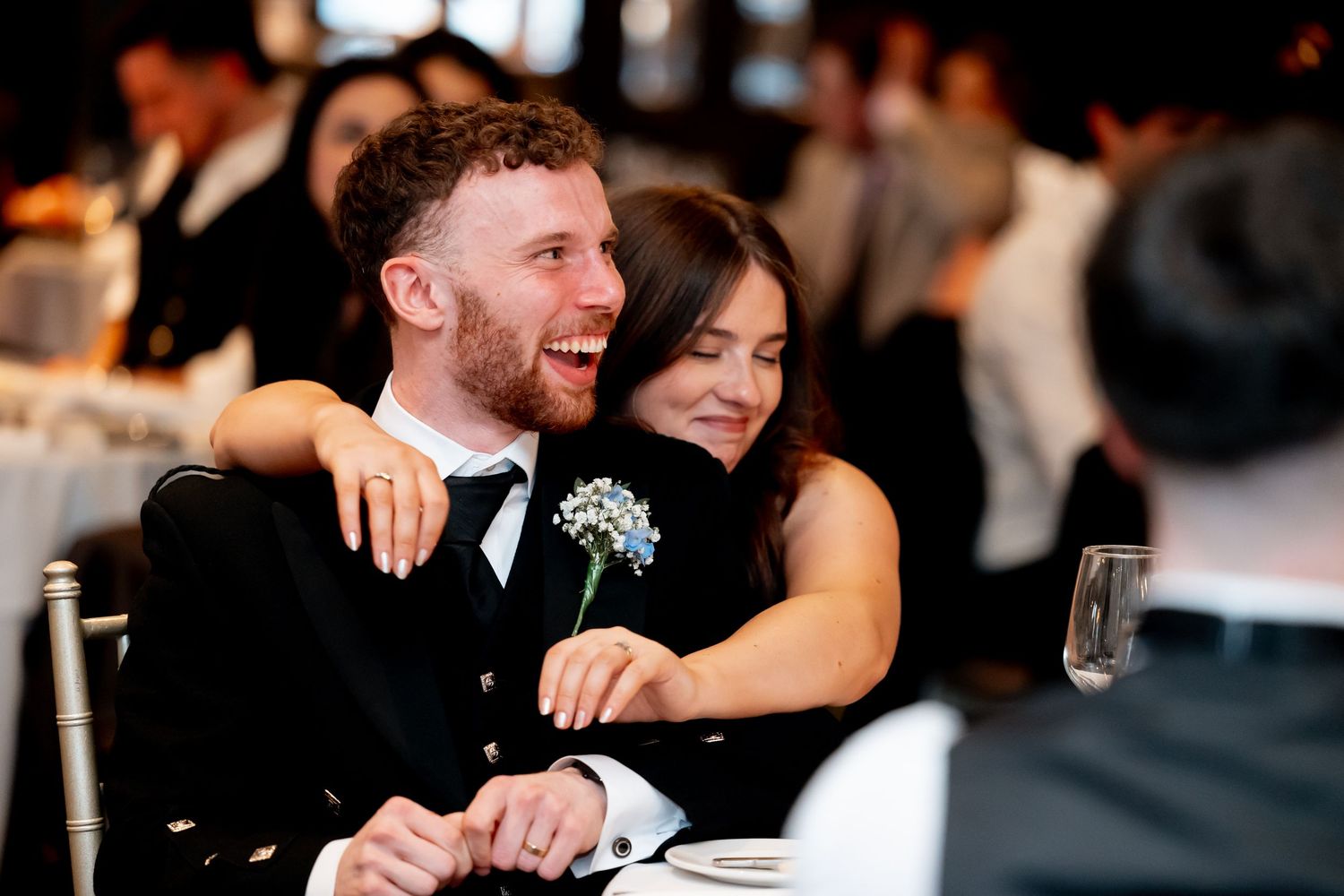 Couple laughing and embracing at wedding reception, groom in black suit with boutonniere, bride hugging him from behind.