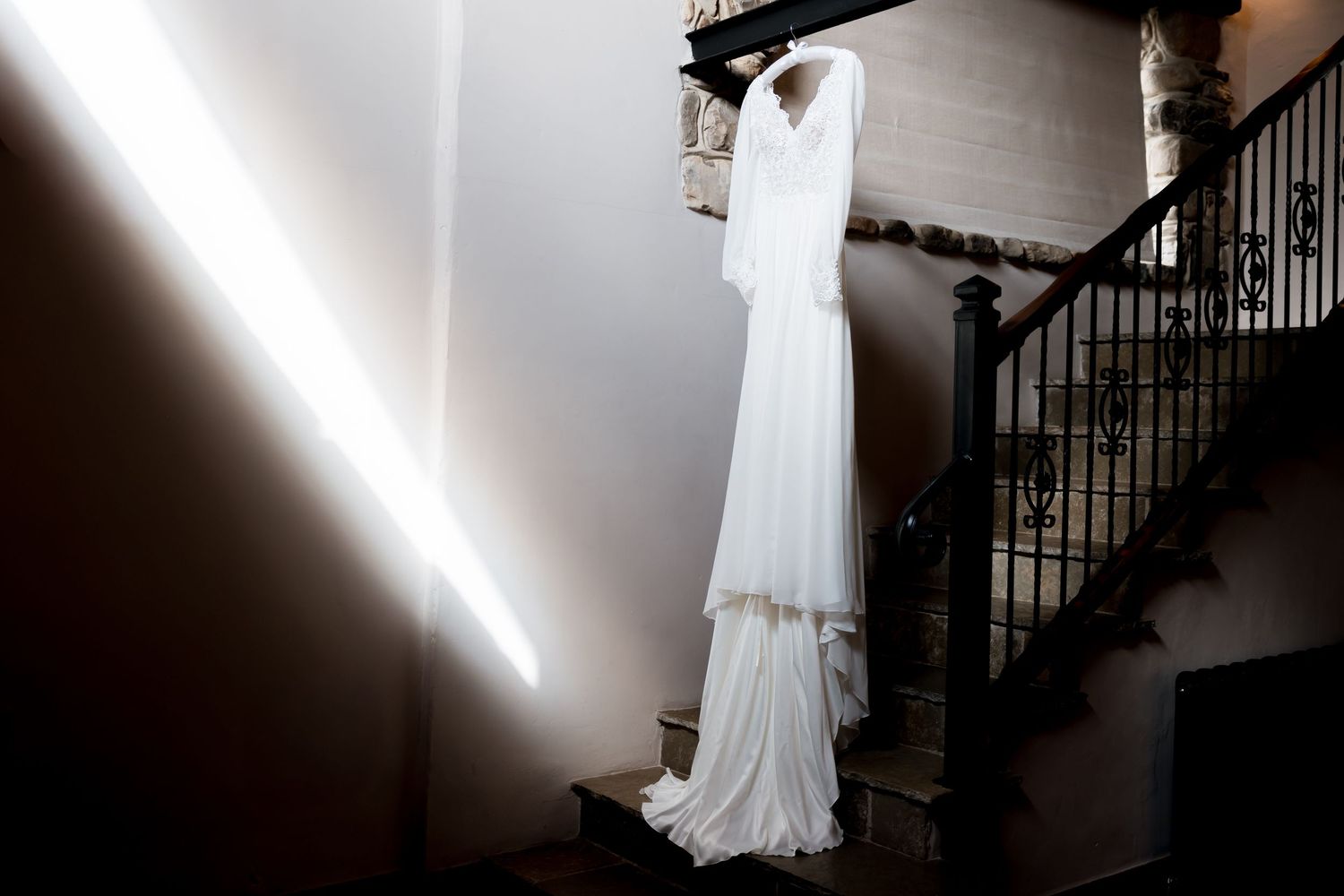 Elegant white wedding dress hanging dramatically in natural light against dark background.