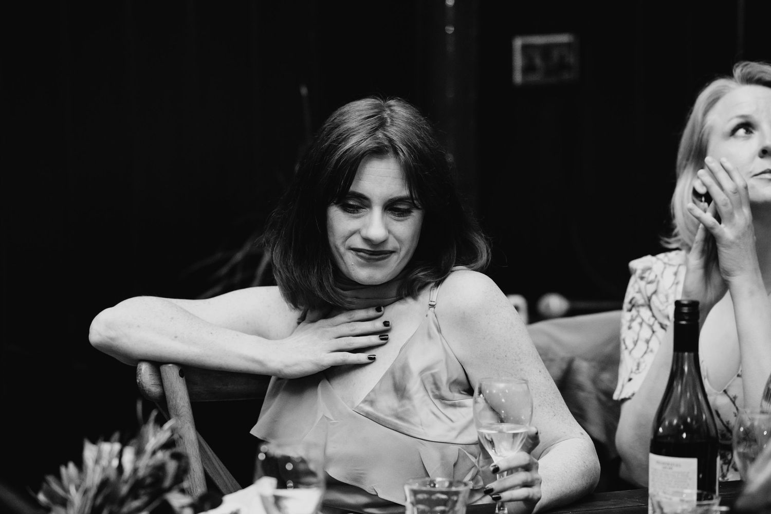 Black and white photo of a woman laughing at a dinner party table with drinks and a wine bottle nearby.