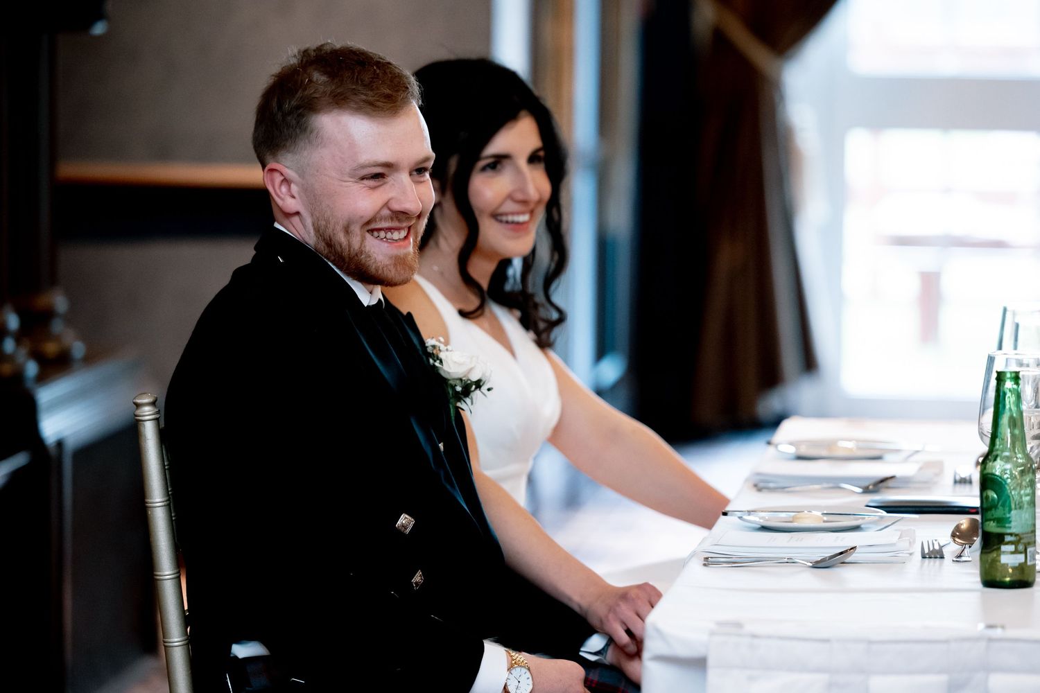 Smiling groom in Scottish kilt jacket and bride in white dress seated together at wedding reception table.