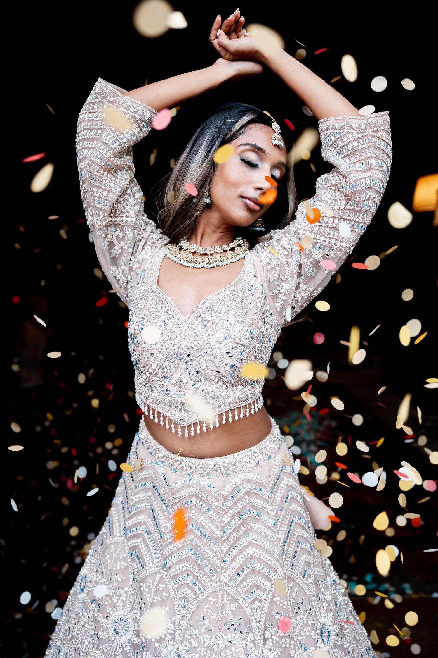 A woman in a silver embroidered lehenga dances gracefully with arms raised as colorful confetti falls around her.