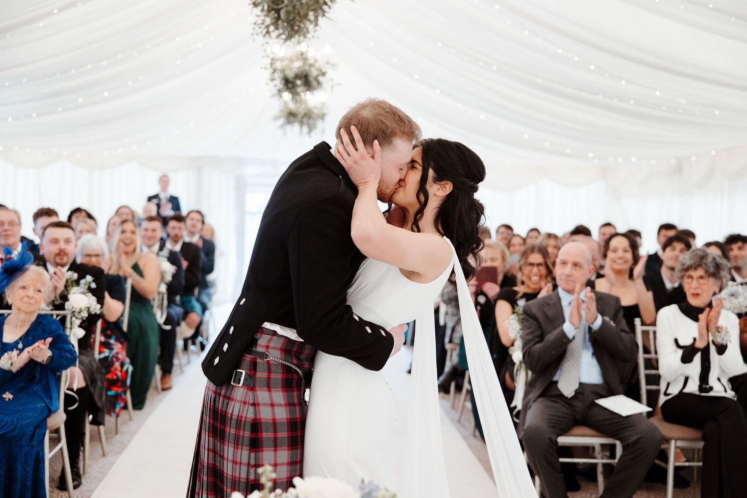 Bride and groom share first kiss at Scottish wedding ceremony, groom wearing traditional tartan kilt, guests applauding in marquee.