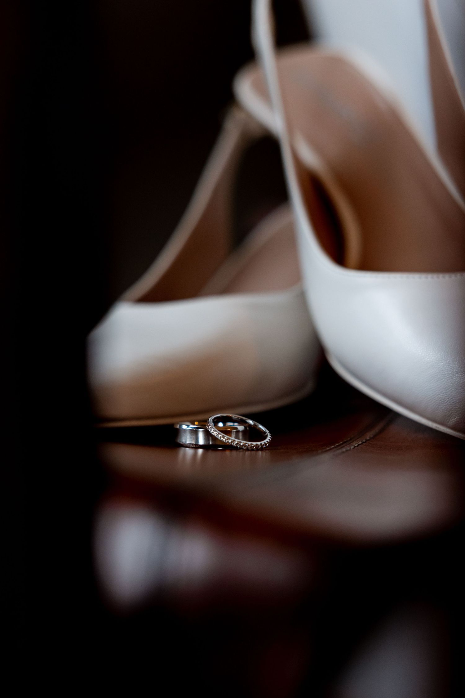 White bridal heels with wedding rings placed in front on a dark reflective surface.