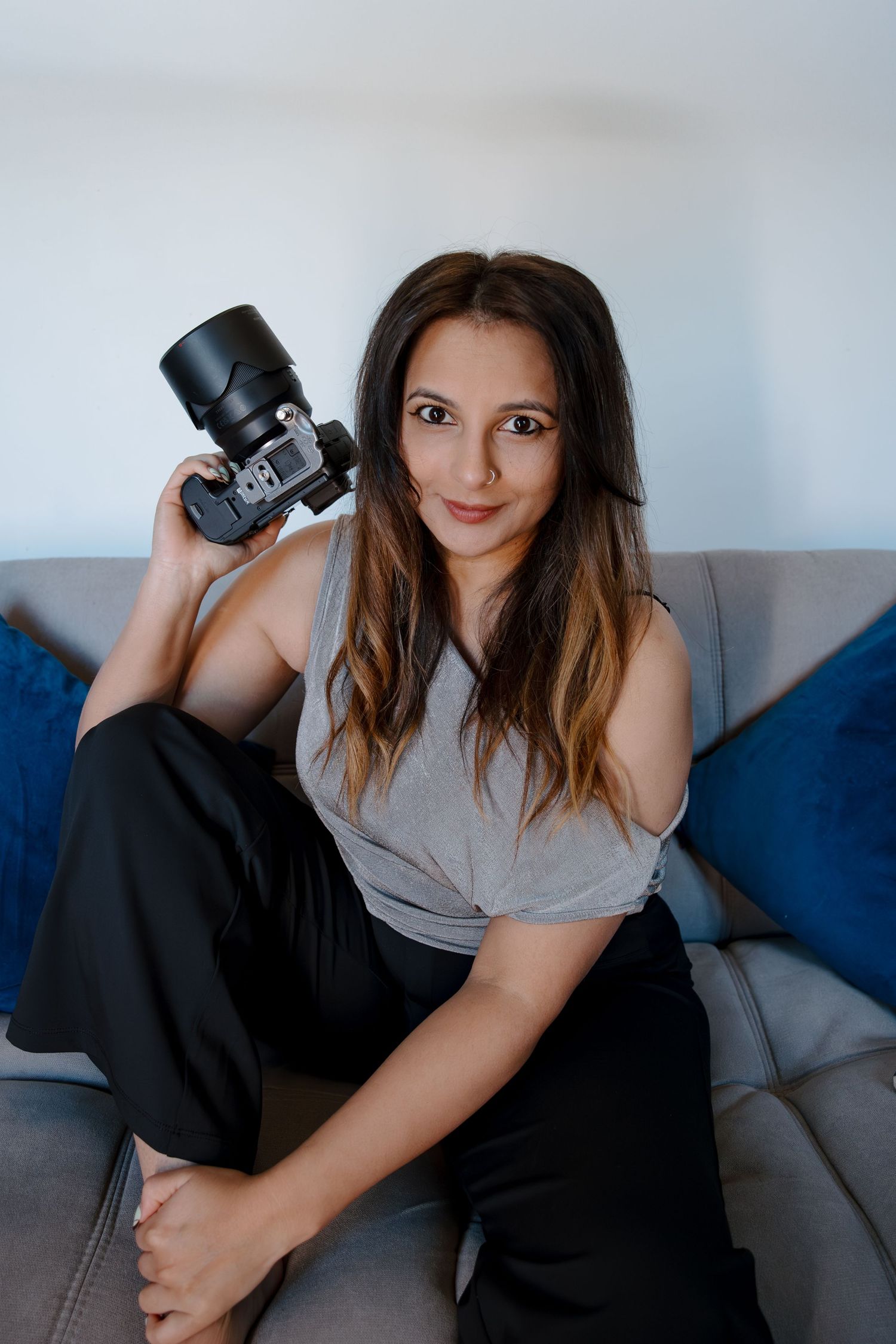 Woman photographer sitting on gray sofa holding DSLR camera, wearing gray top and black pants.