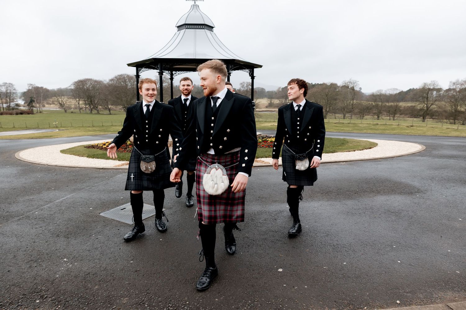 Four men in traditional Scottish kilts and black jackets walk near a bandstand in a park.