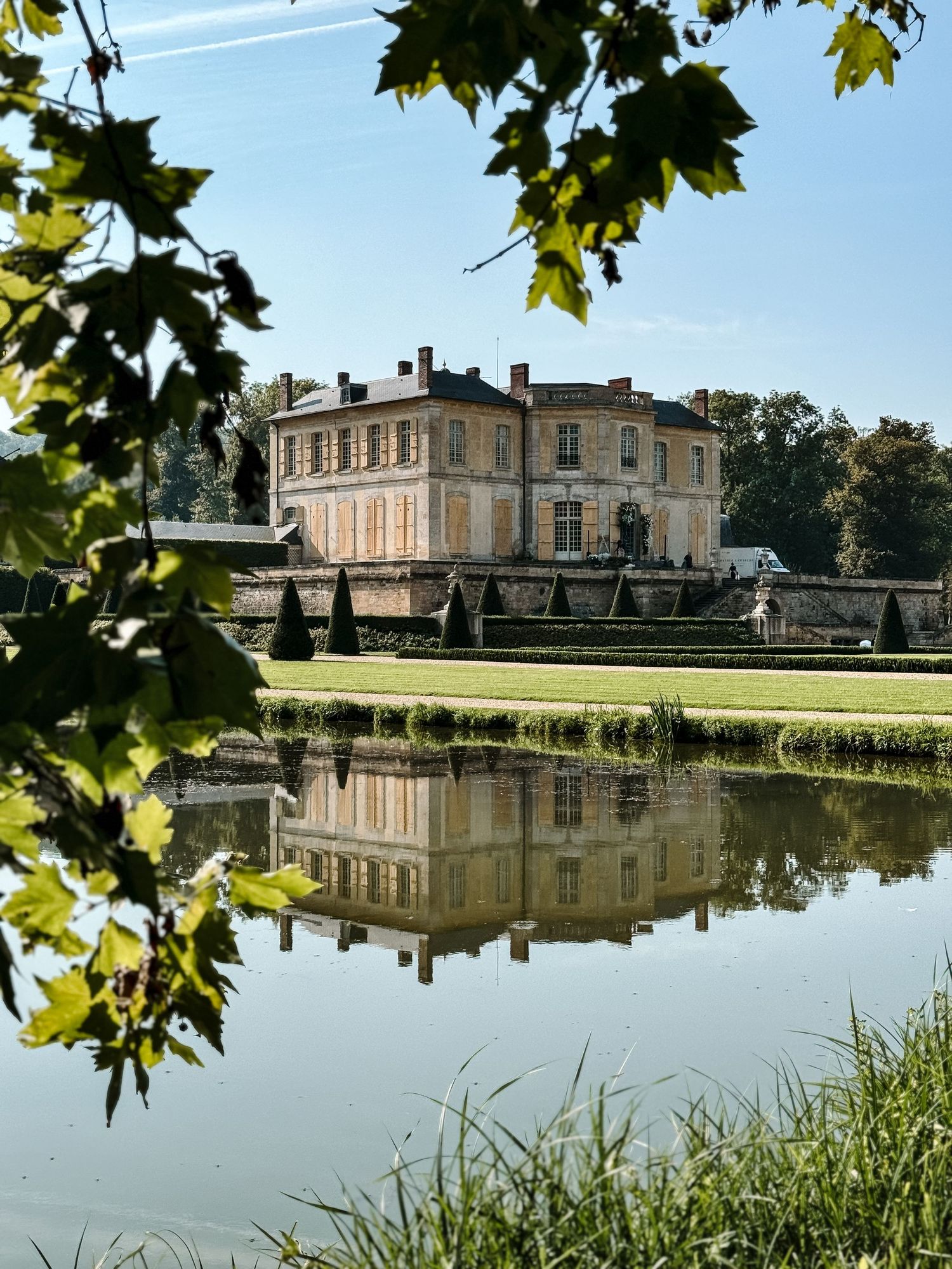 Grand mansion reflected in still lake waters with manicured gardens and maple leaves framing the view.