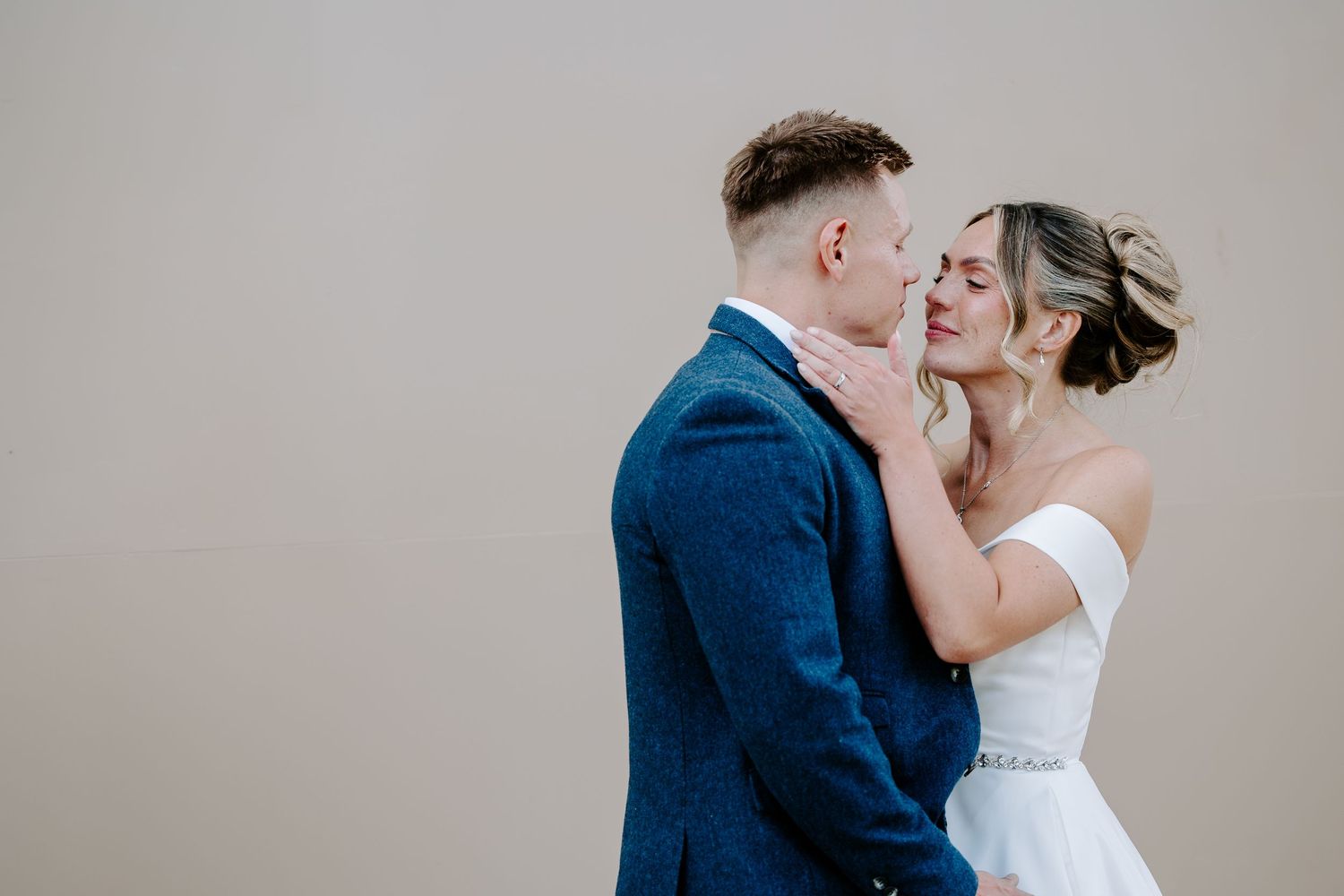 A couple shares a romantic moment in formal wedding attire against a neutral background.