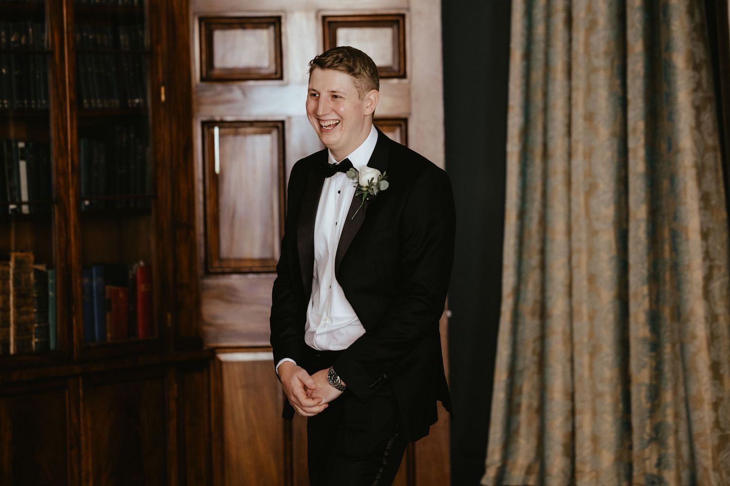 Smiling groom in black tuxedo with white boutonniere laughing joyfully in elegant wood-paneled room.