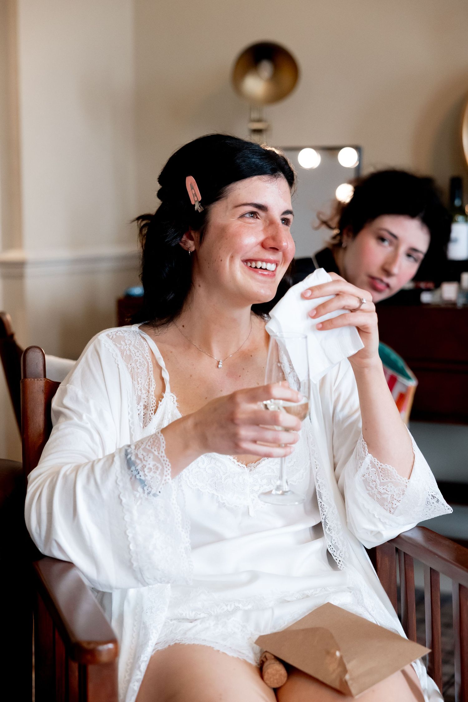 Bride in white lace robe smiling and holding champagne glass while getting ready on wedding day.