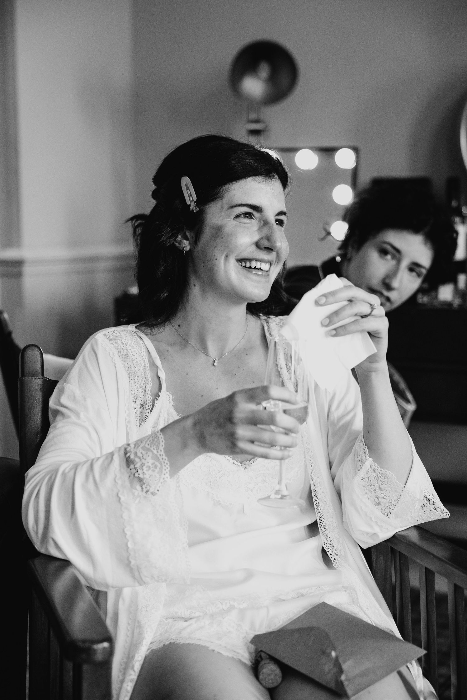 Black and white photo of a smiling bride in a lace robe holding a champagne glass while getting ready.