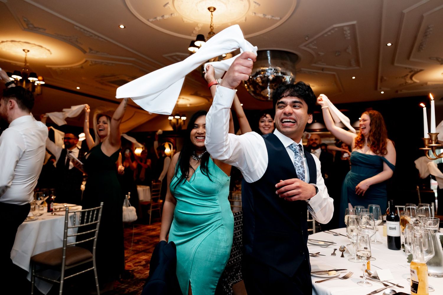 Guests celebrate at a wedding reception, waving napkins and dancing joyfully in an elegant banquet hall.
