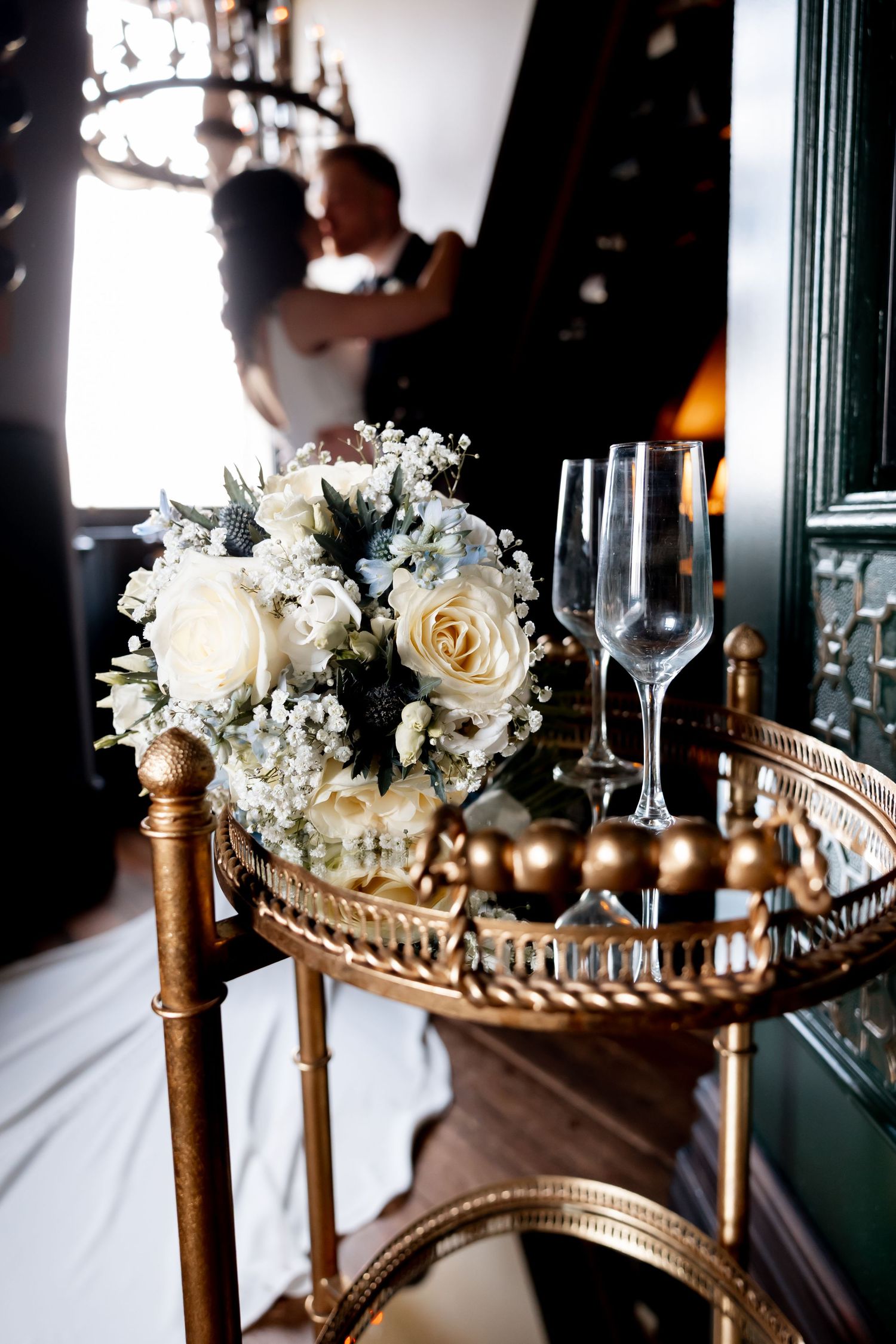 White rose bridal bouquet with champagne glasses on gold tray, couple embracing in background.