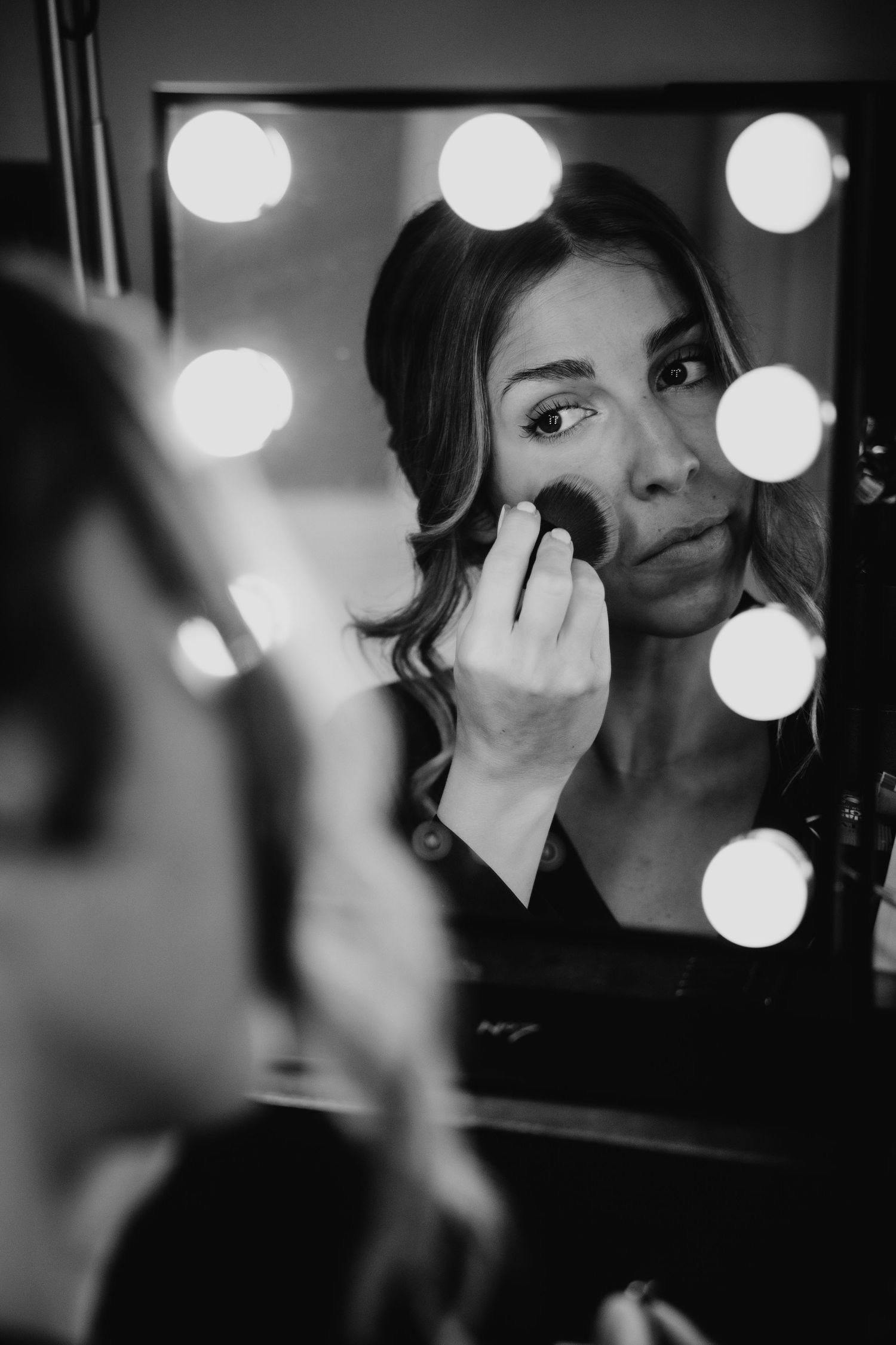 Woman applying blush with makeup brush in front of Hollywood vanity mirror, black and white photo.