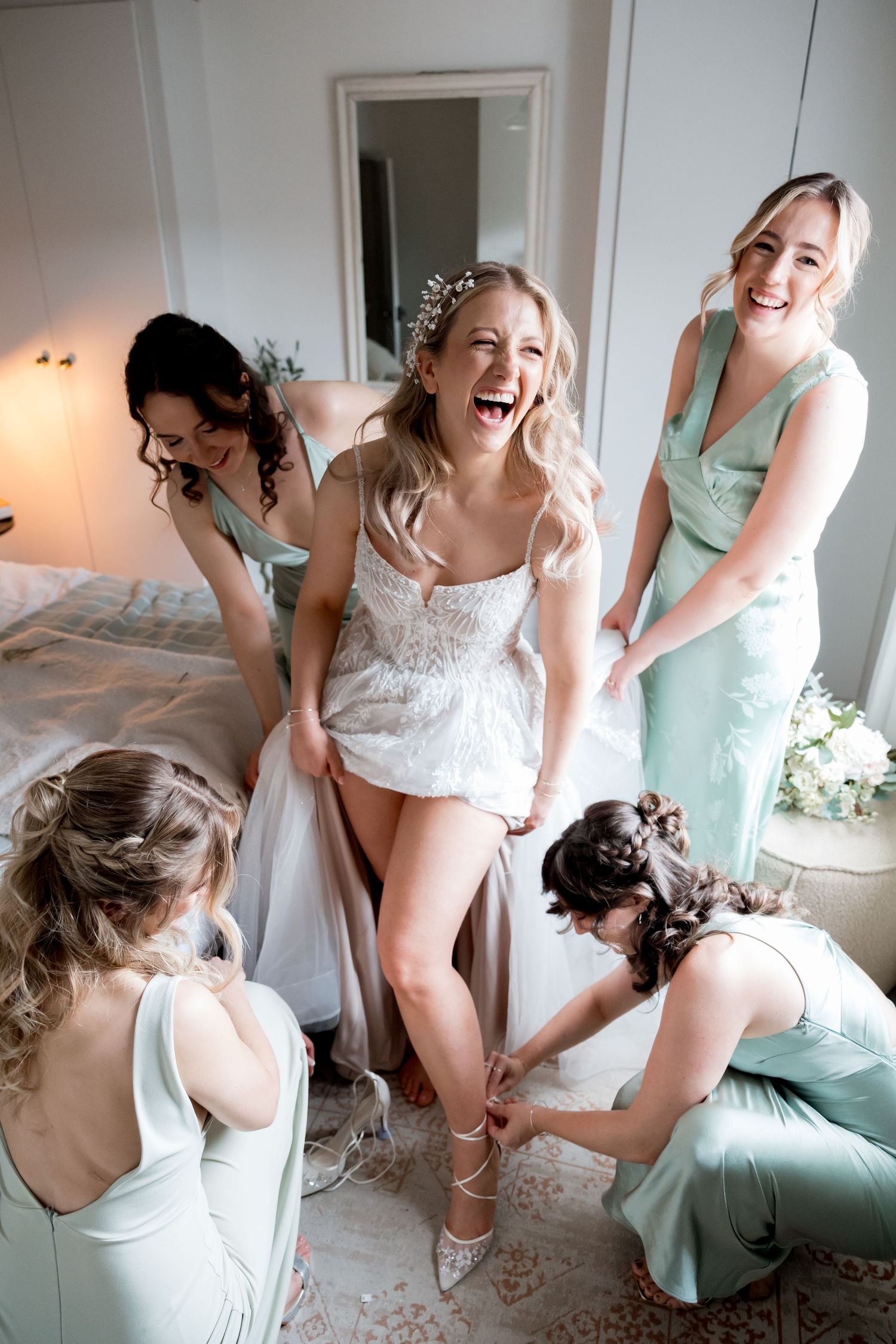Bride laughing joyfully with bridesmaids in mint dresses helping her get ready, fastening shoes in bright bedroom.