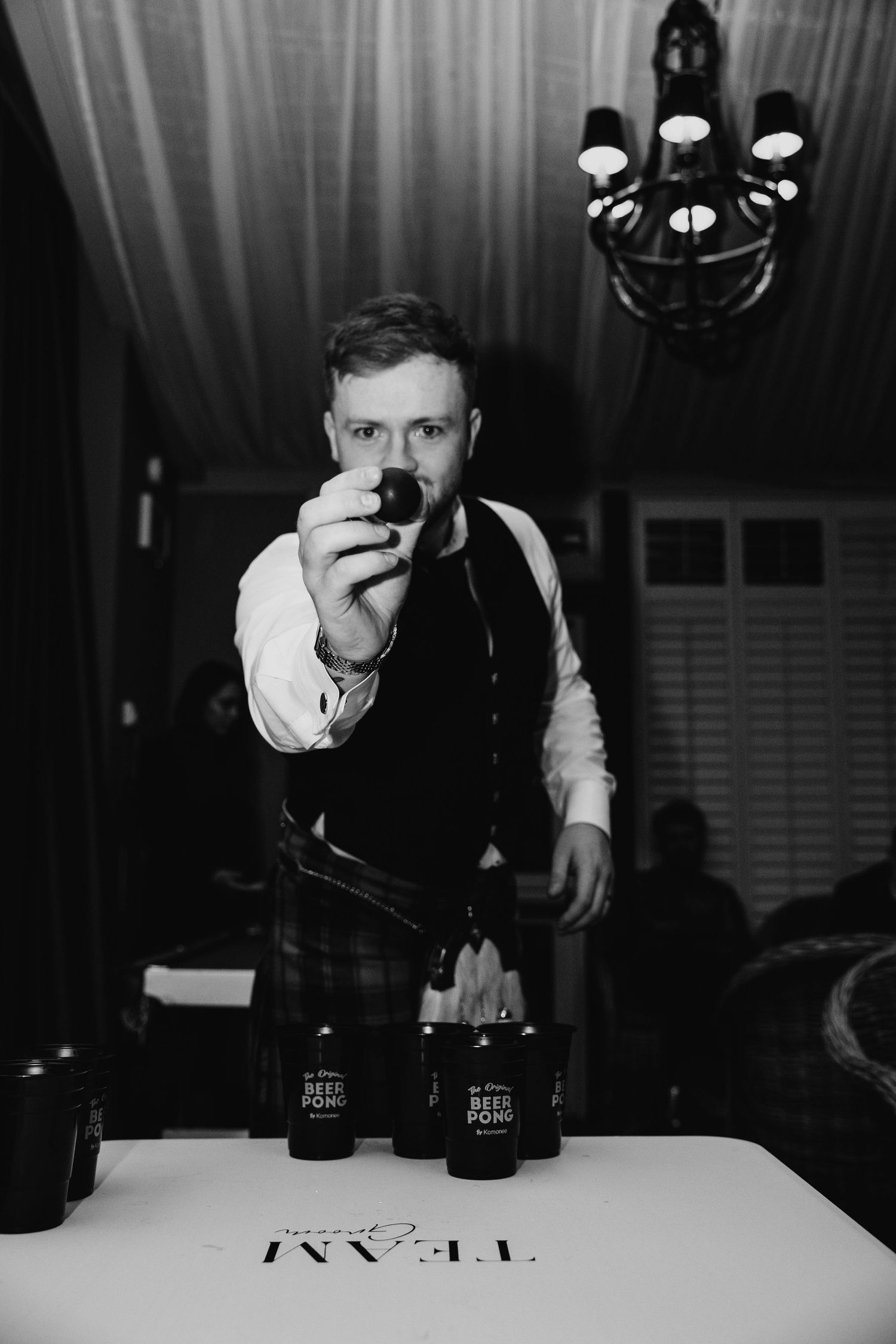 Man holding ping pong ball over beer pong table with cups at indoor party event, black and white photo.