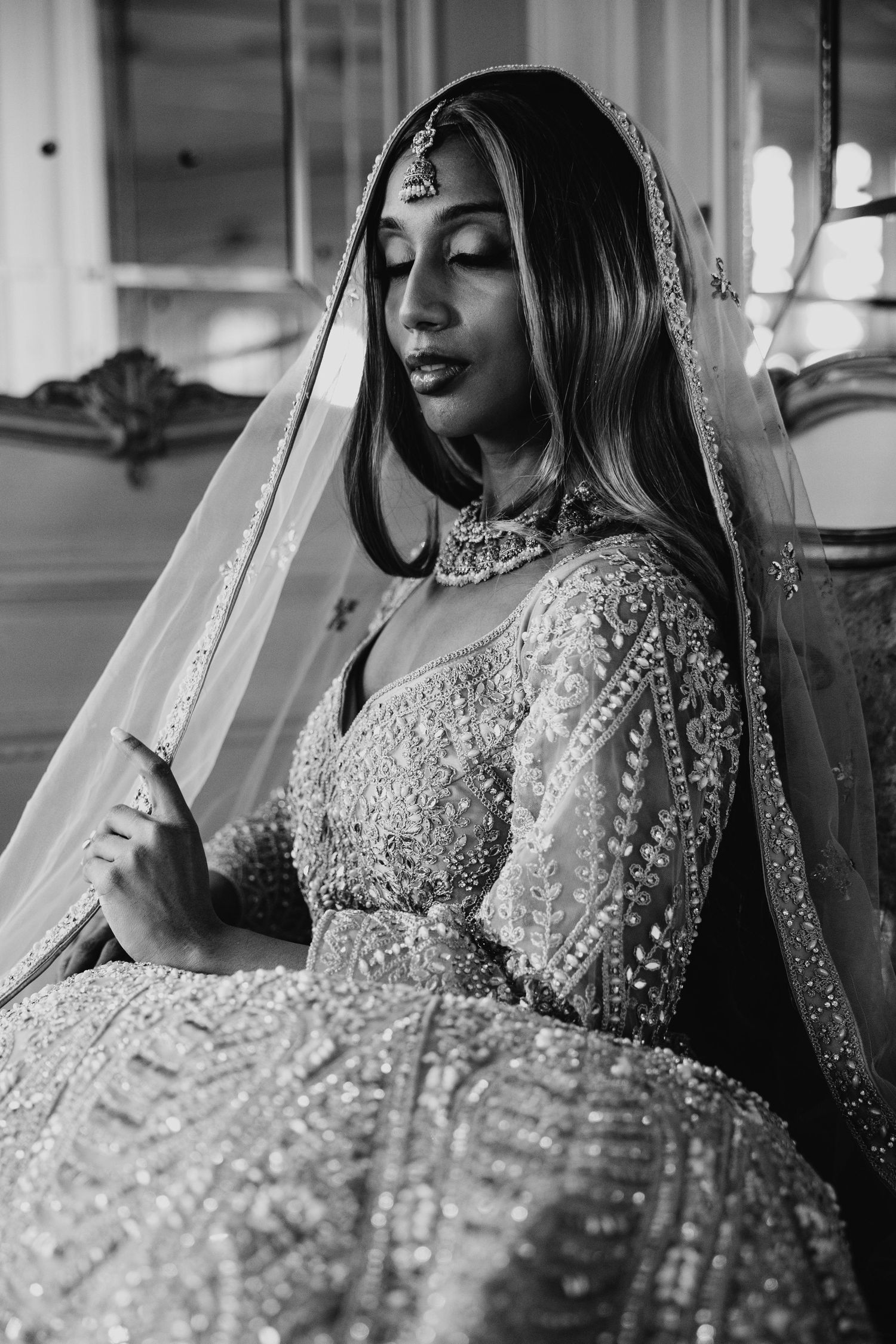 Black and white portrait of a bride in ornate Indian bridal lehenga with embroidery, dupatta veil, and maang tikka jewelry.