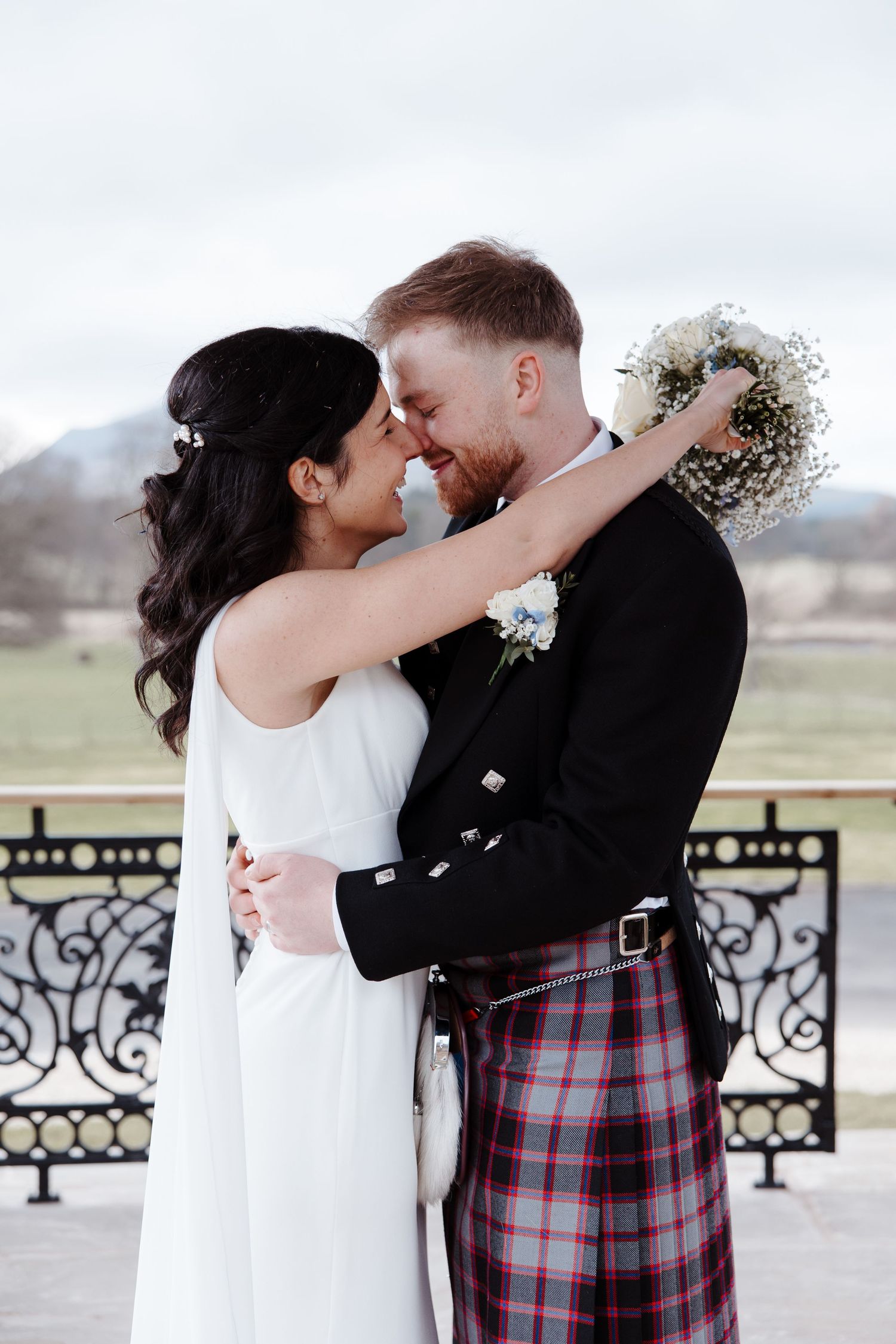 A bride and groom share a romantic moment, the groom wearing a traditional Scottish tartan kilt, outdoors at their wedding.