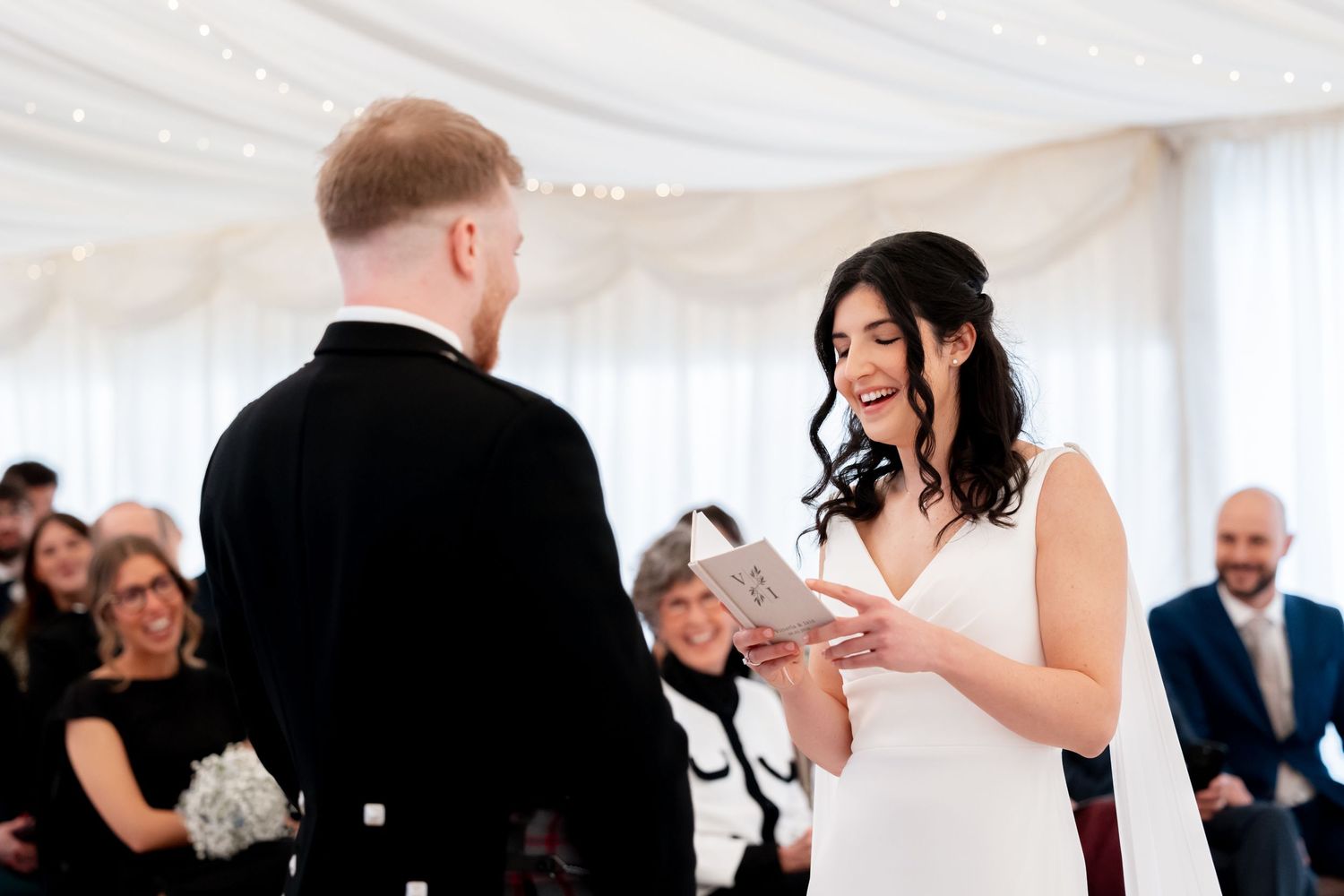 Bride smiling as she places a wedding ring on the groom's finger during a ceremony in a white marquee.