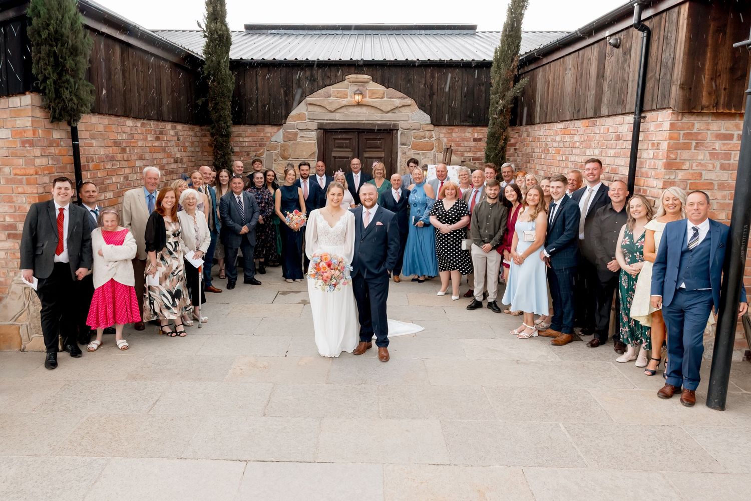 Panoramic shot of wedding guests lined up outside rustic venue.