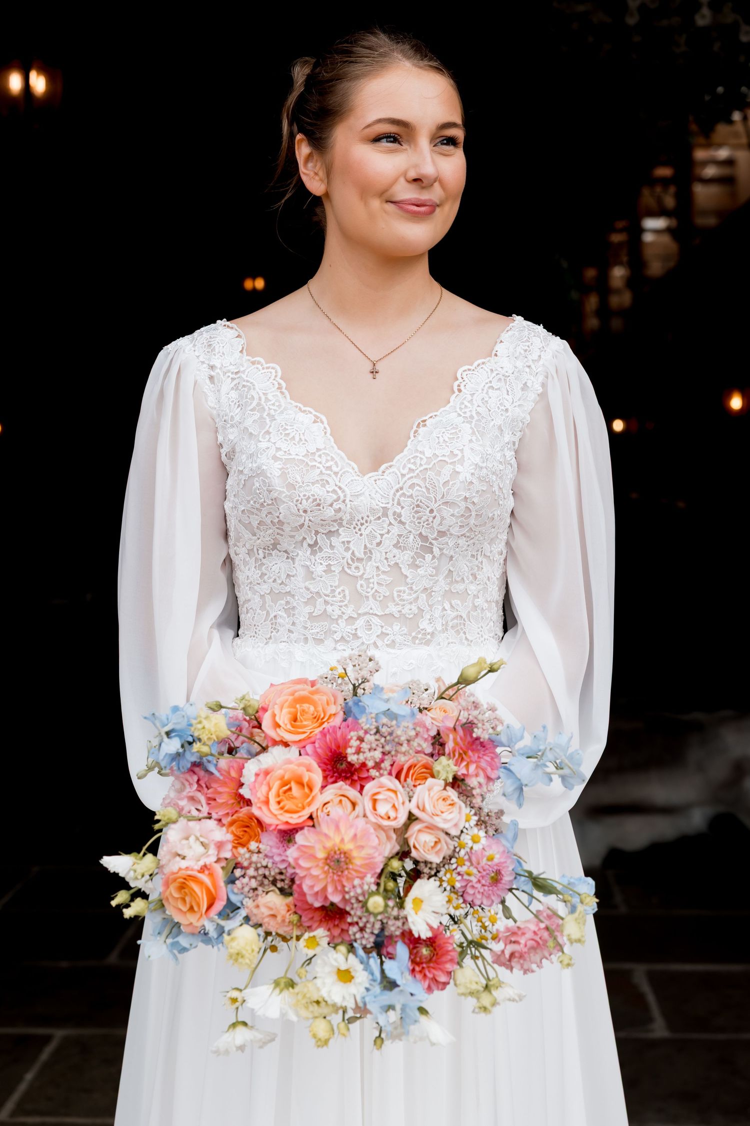 A bride in a white lace dress holds a vibrant bouquet of coral, pink and blue flowers.