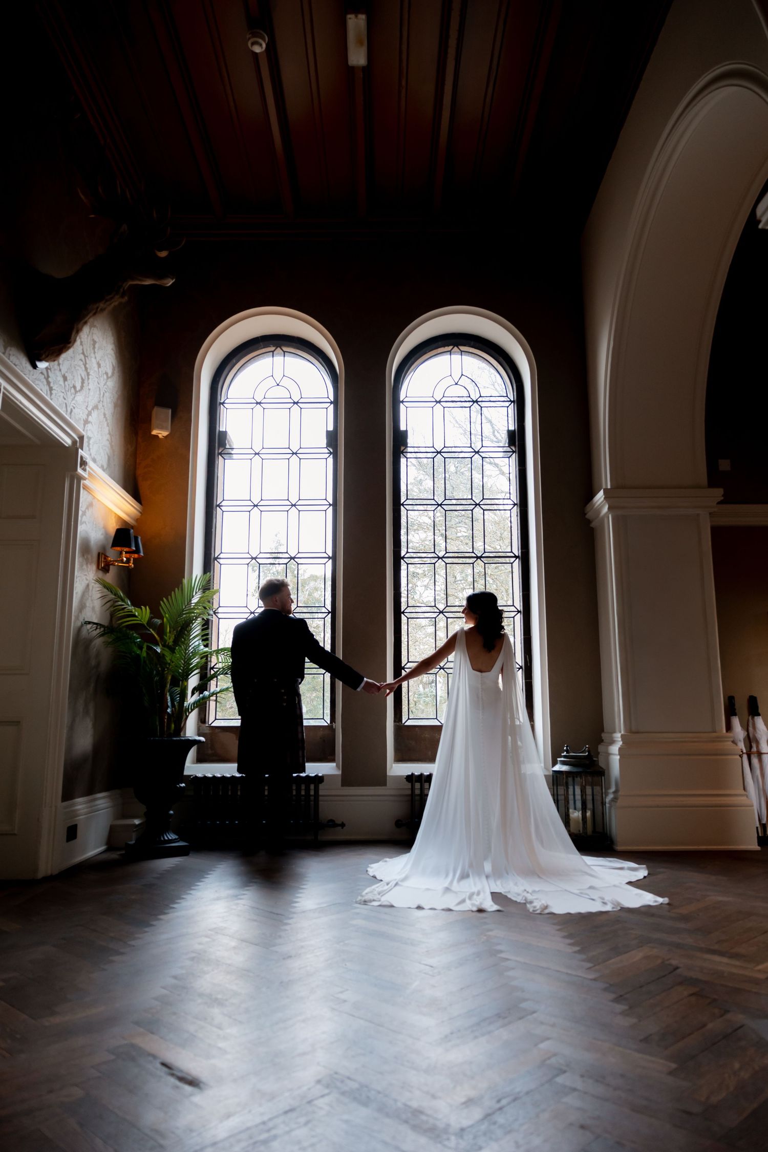 Bride and groom hold hands silhouetted before tall arched stained glass windows in an elegant wedding venue.