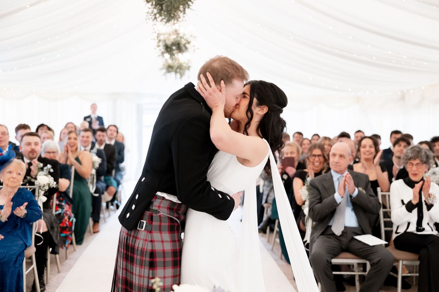 Bride and groom sharing first kiss in white marquee wedding ceremony with guests applauding.