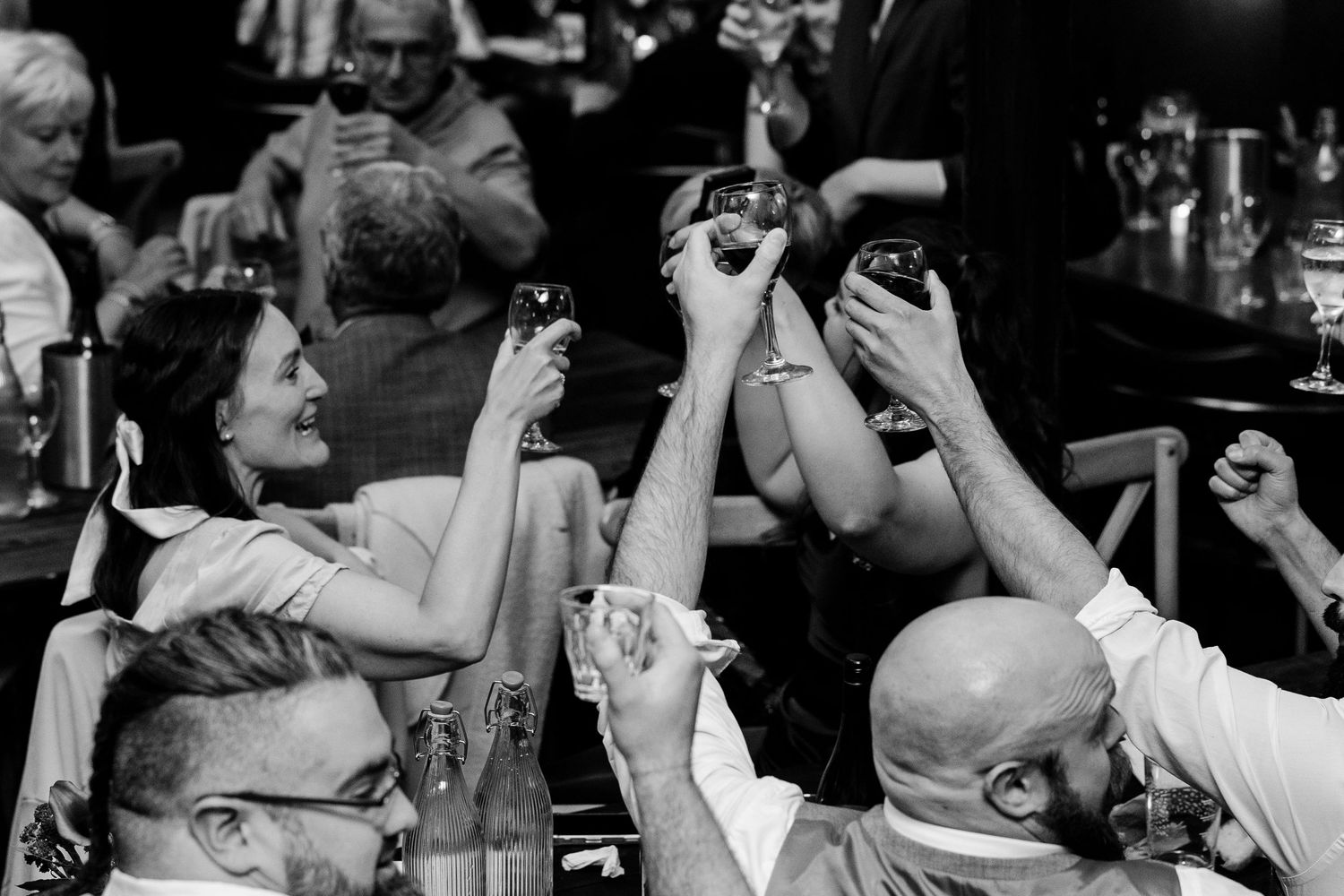 Black and white photo of wedding guests raising glasses in a joyful toast at a reception celebration.