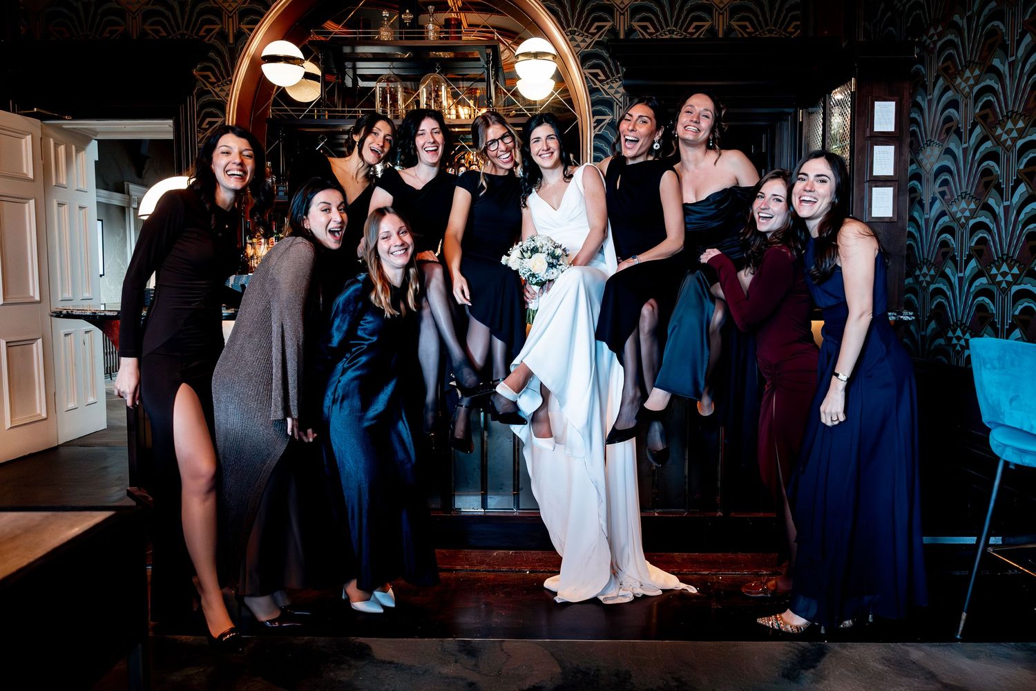 Bride poses with bridesmaids in elegant dresses, laughing together in a stylish dark-walled venue.
