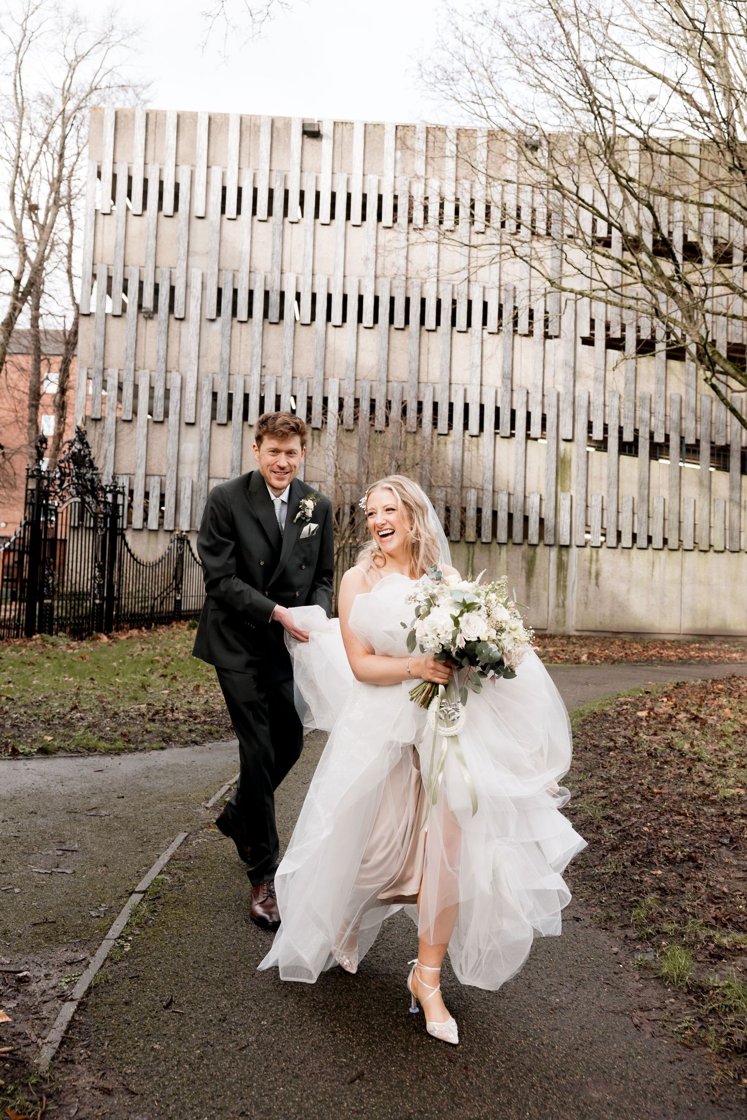 A joyful bride in a white gown holding a bouquet walks with a groom in a black suit on a paved path outdoors.