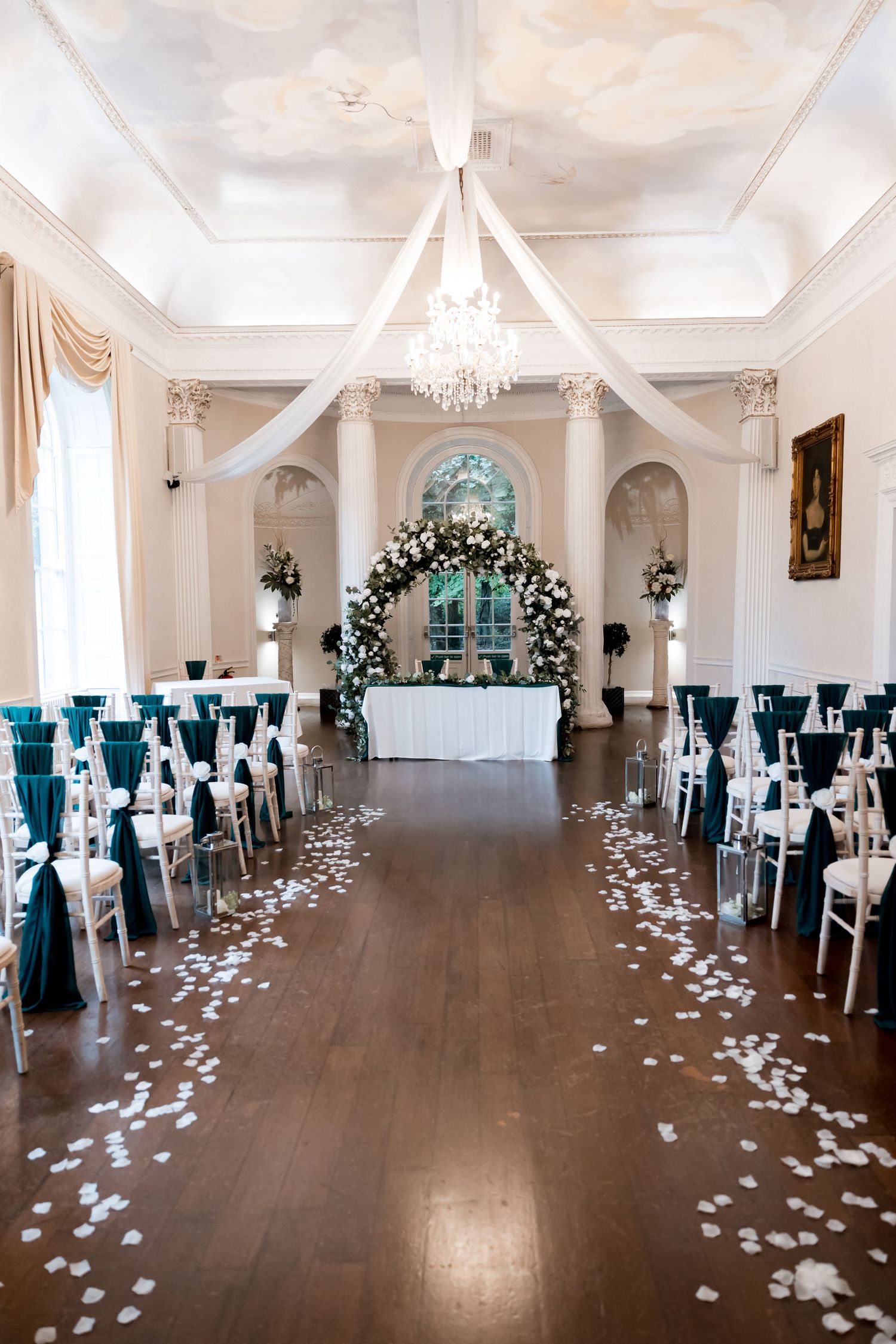 Elegant white ballroom with teal chair sashes and scattered rose petals on hardwood floor.