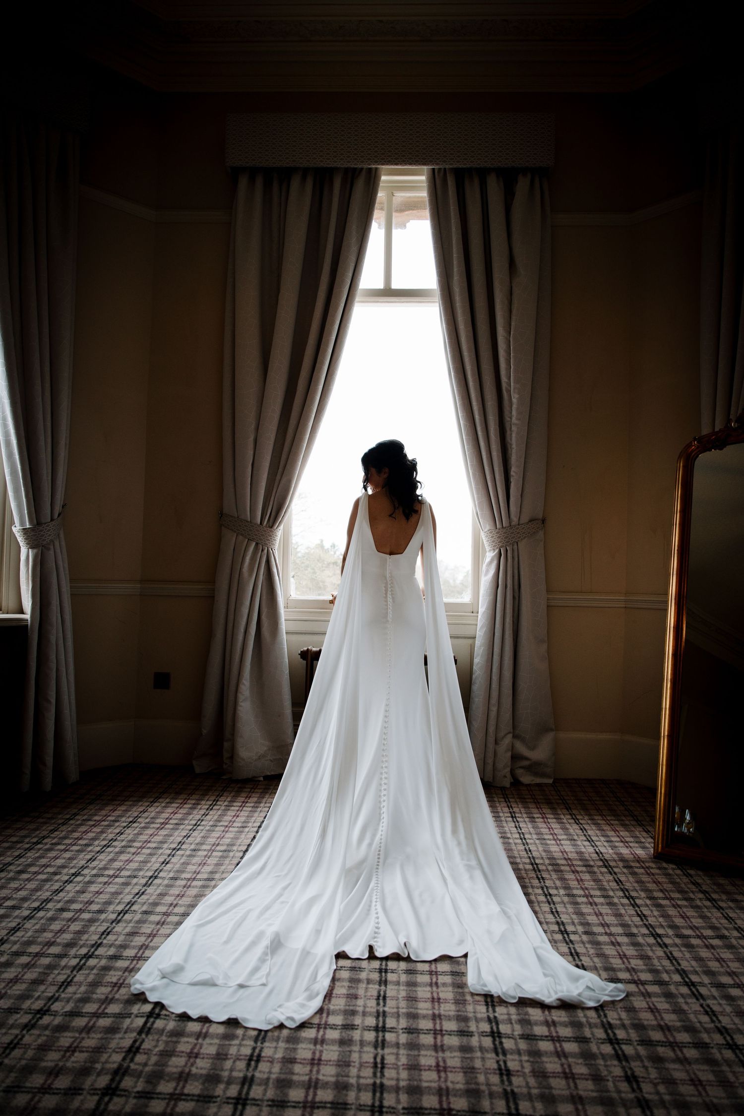 Bride in elegant white gown with long train and flowing sleeves standing before a window in a dimly lit room.