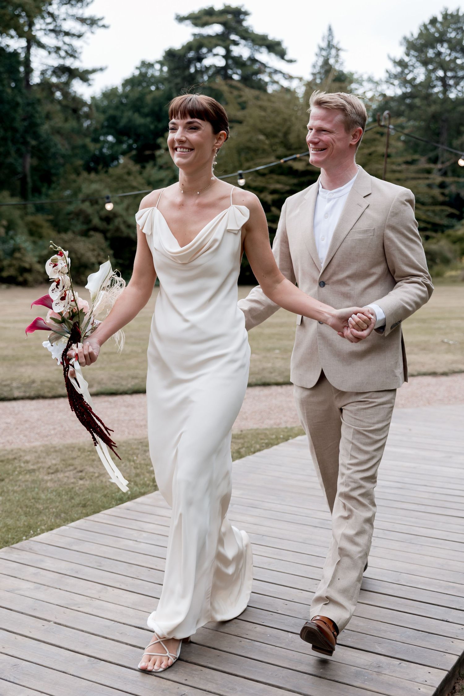 Couple walking hand in hand on wooden deck in wedding attire.