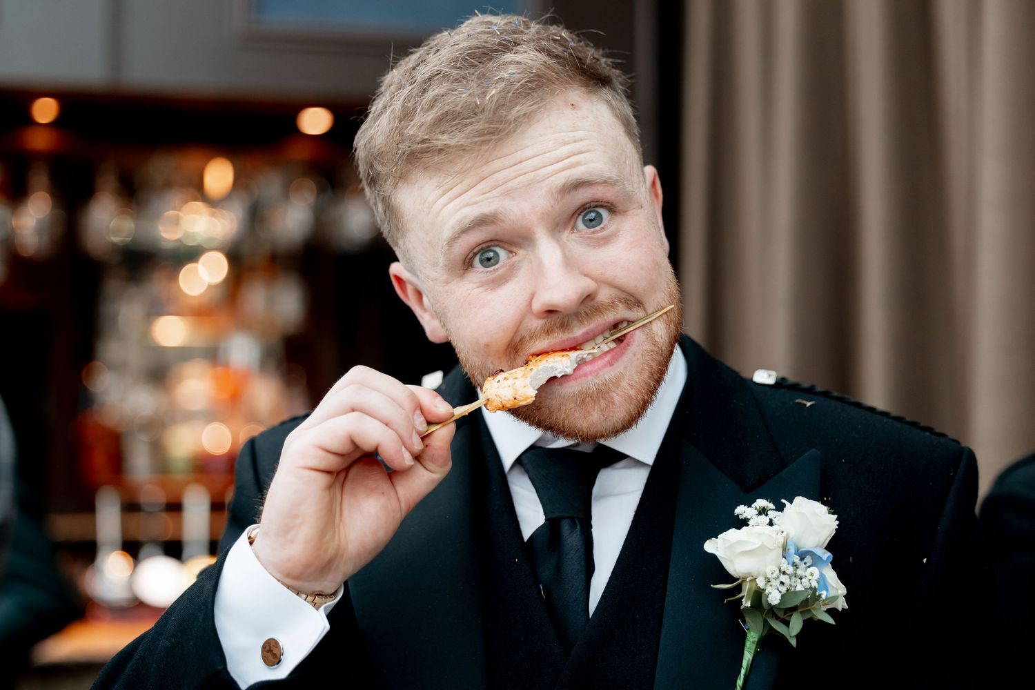 Smiling groom in black suit with boutonniere playfully holding appetizer at wedding reception.