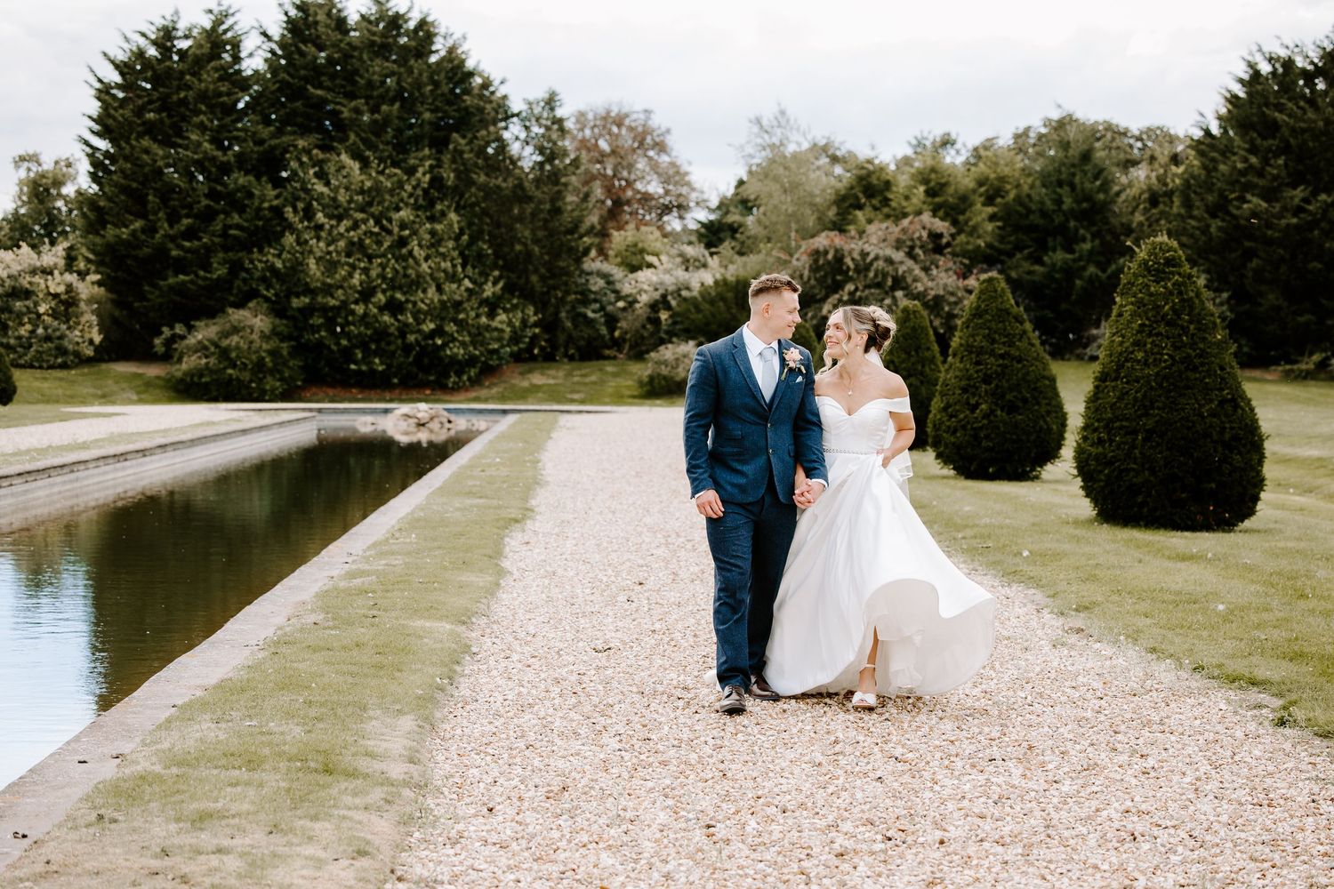 A couple walks along a scenic gravel path beside a tranquil lake on a sunny day.