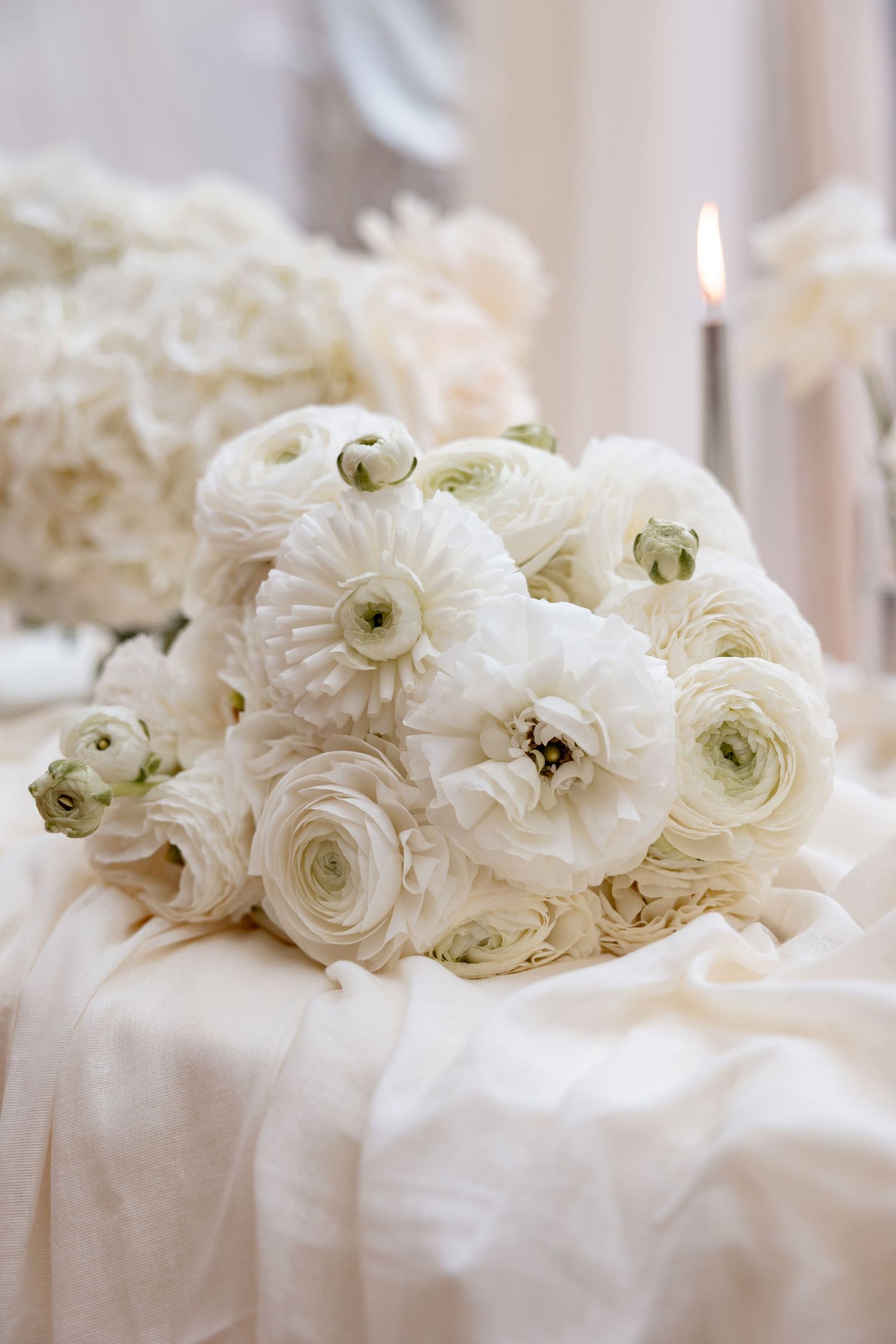 Elegant white ranunculus bouquet with candle on draped fabric at a wedding reception.
