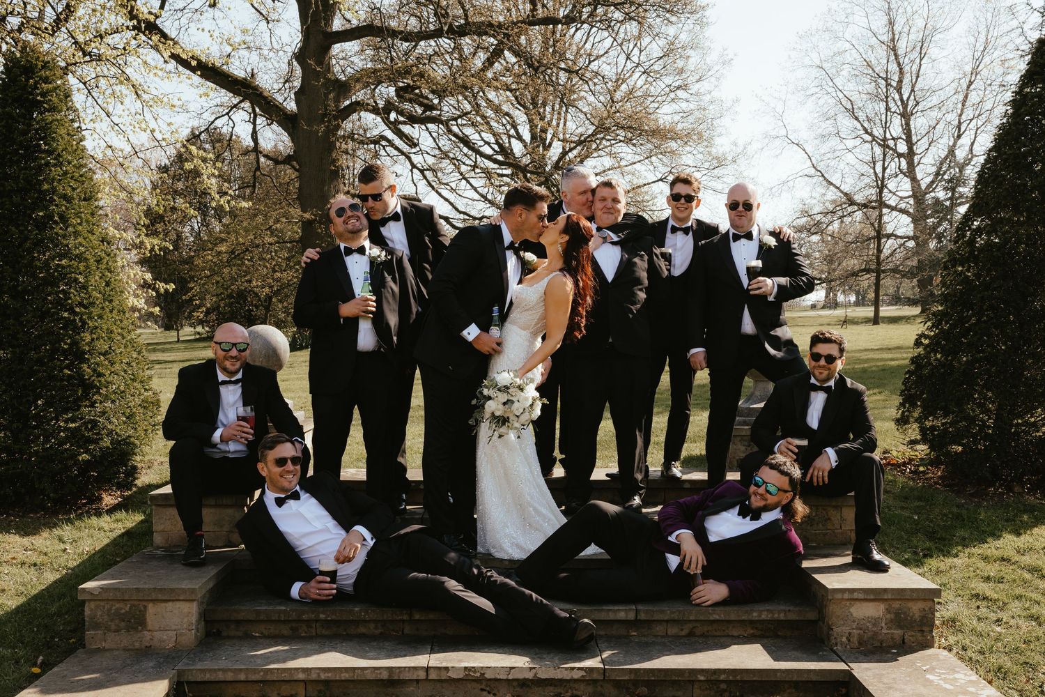 Wedding party photo with bride and groom kissing surrounded by groomsmen in black tuxedos on outdoor stone steps.