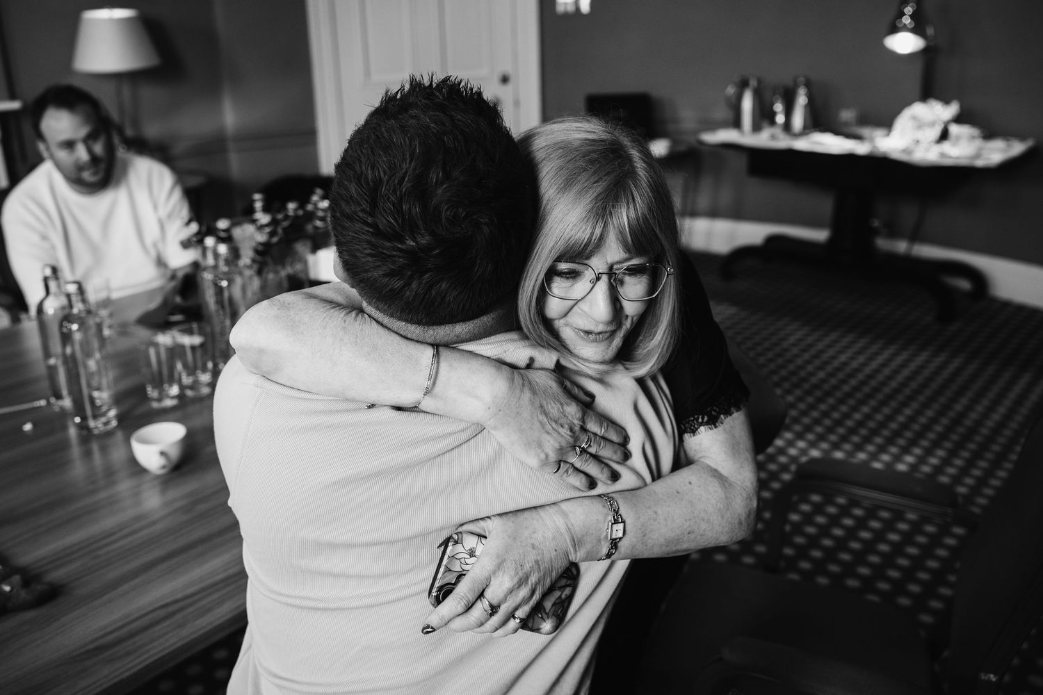 Black and white photo of two people sharing a warm hug in a restaurant, with a man seated at a table in the background.