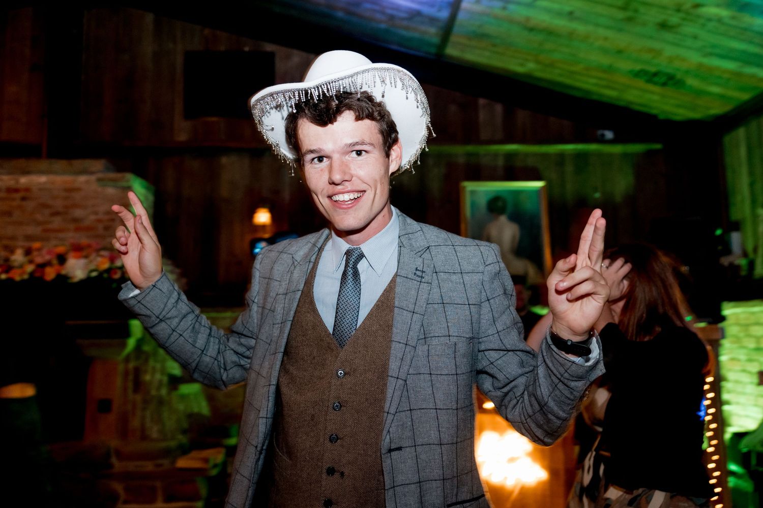 Party guest in grey suit and cowboy hat making peace signs while dancing.