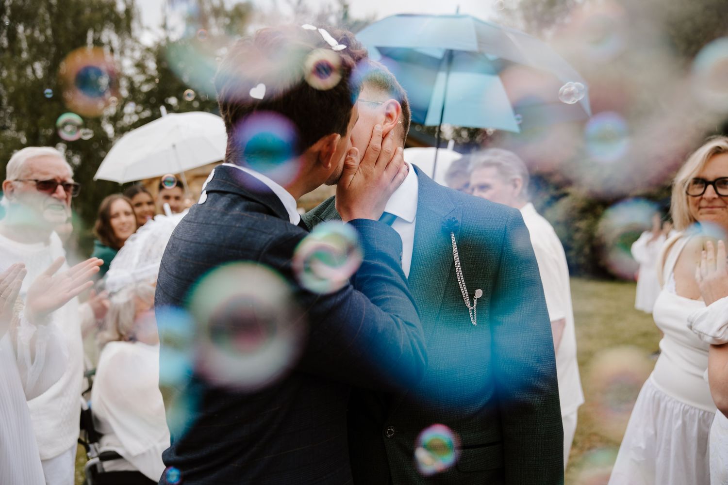 Wedding guests hold blue umbrellas and laugh as bubbles float through the air during an outdoor celebration.