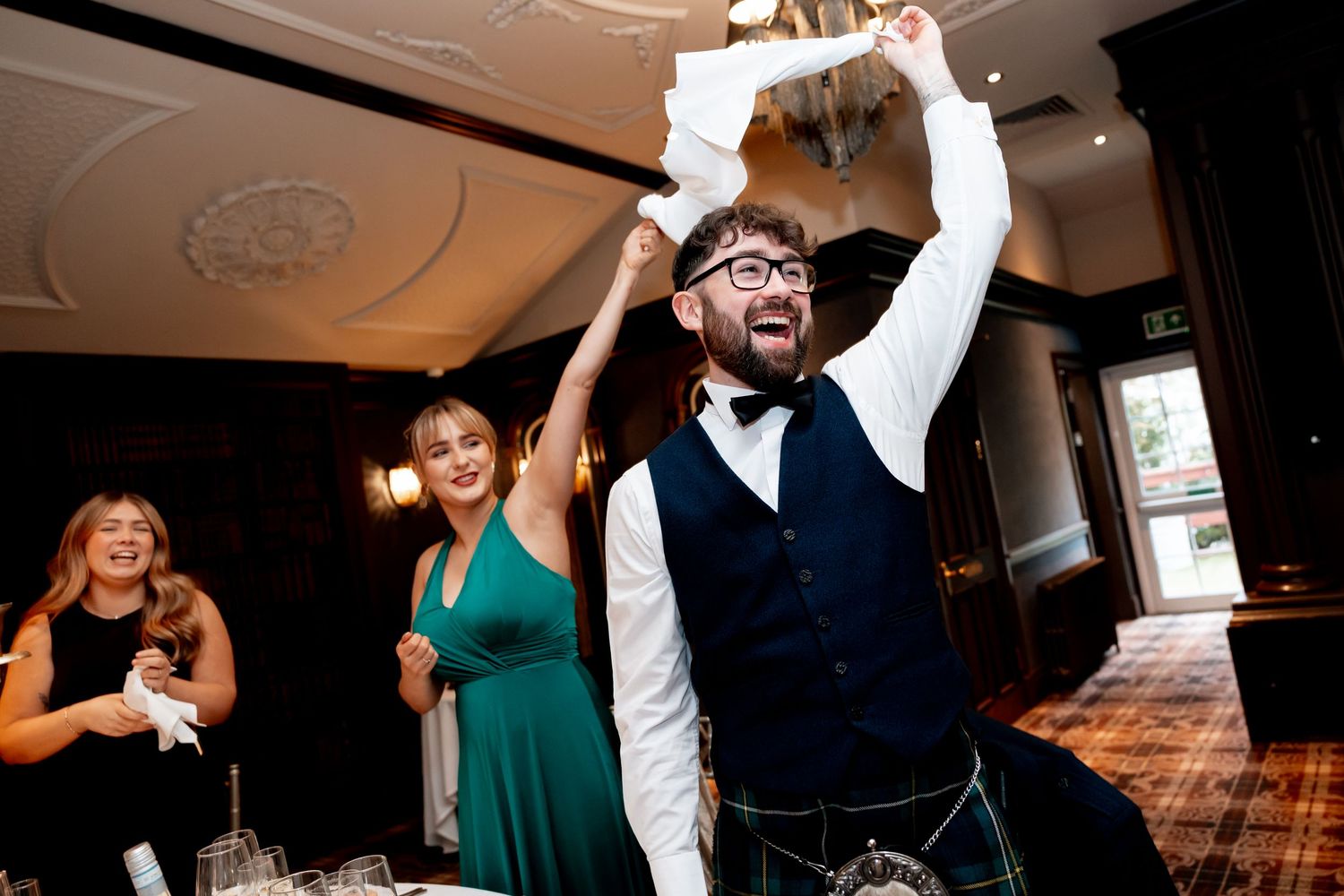 Excited wedding guest in a kilt raising papers in the air during a lively reception celebration.