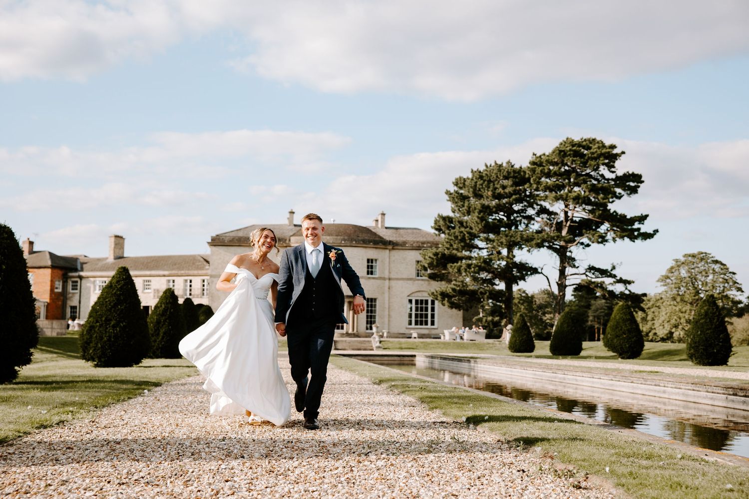 A wedding couple walks along a scenic coastal path with elegant buildings and landscaping in the background.