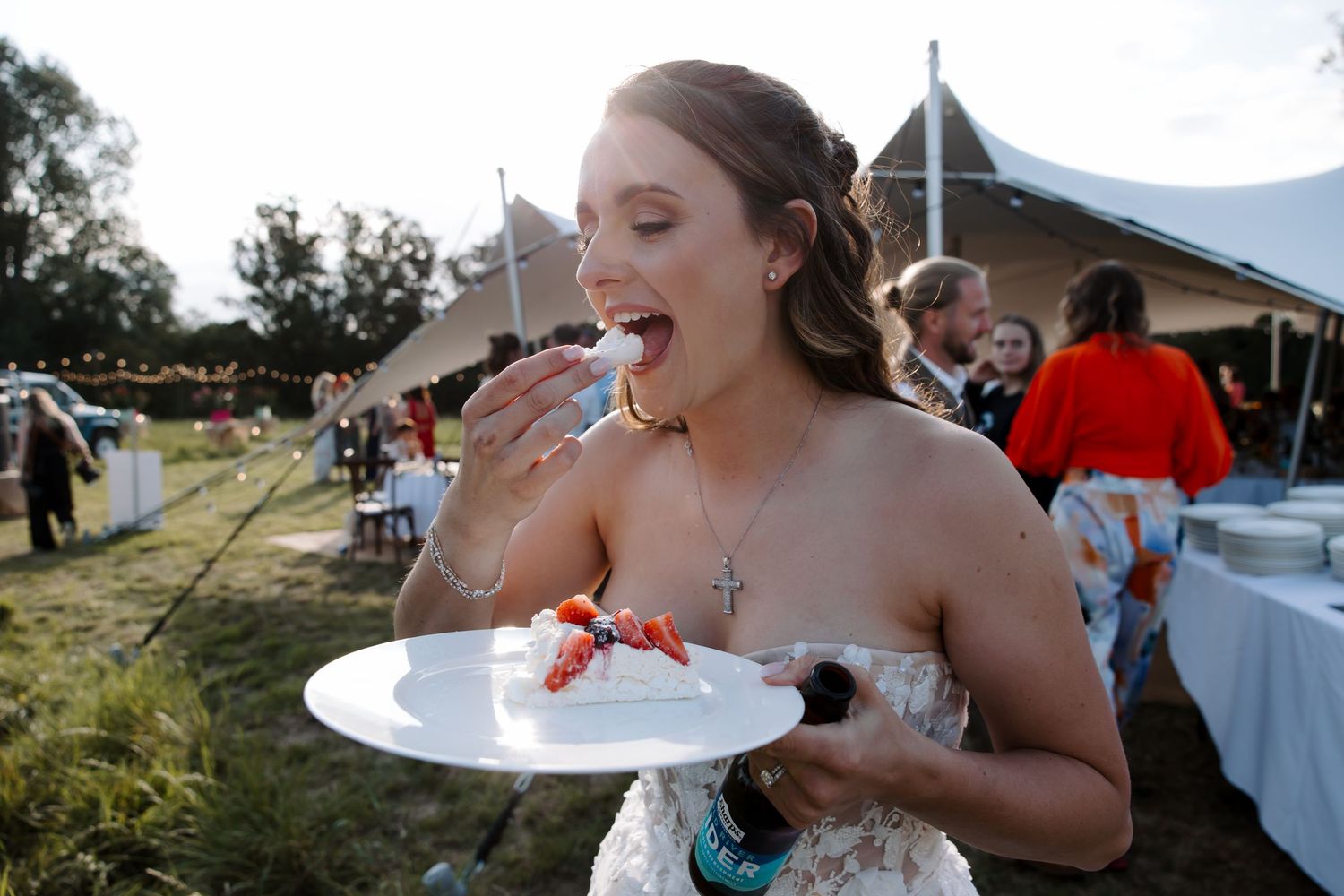 A wedding guest enjoys cake at an outdoor reception under a tent.