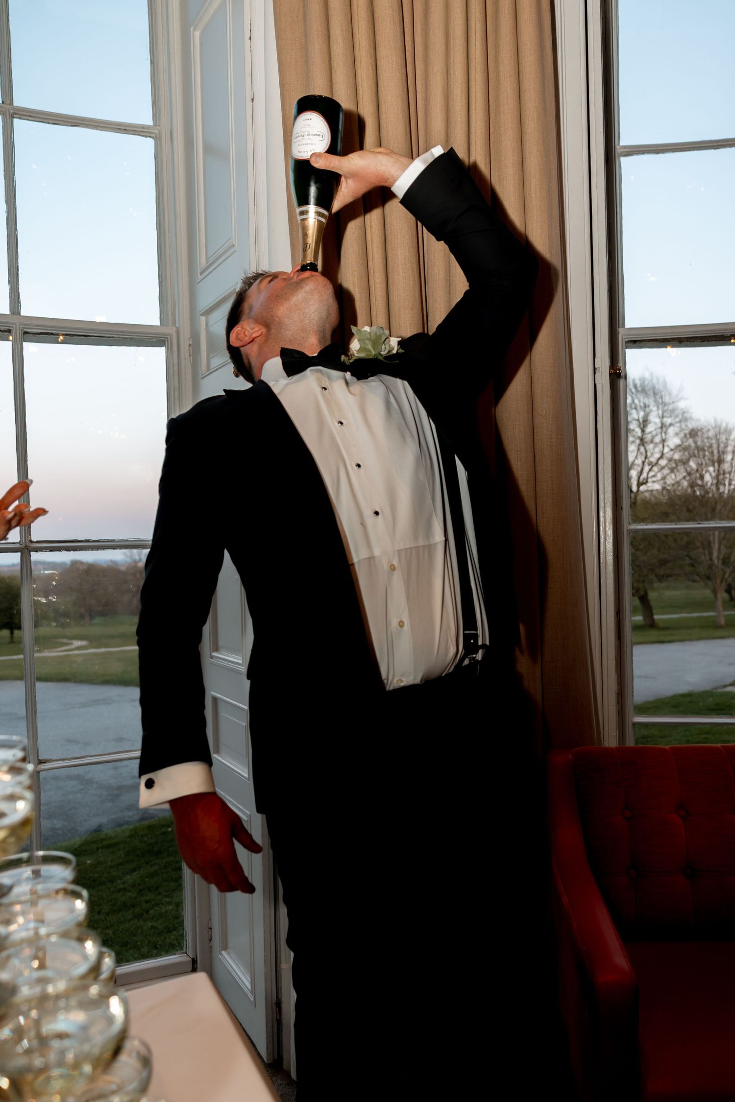 Groom in tuxedo drinking from champagne bottle at wedding reception near large windows.