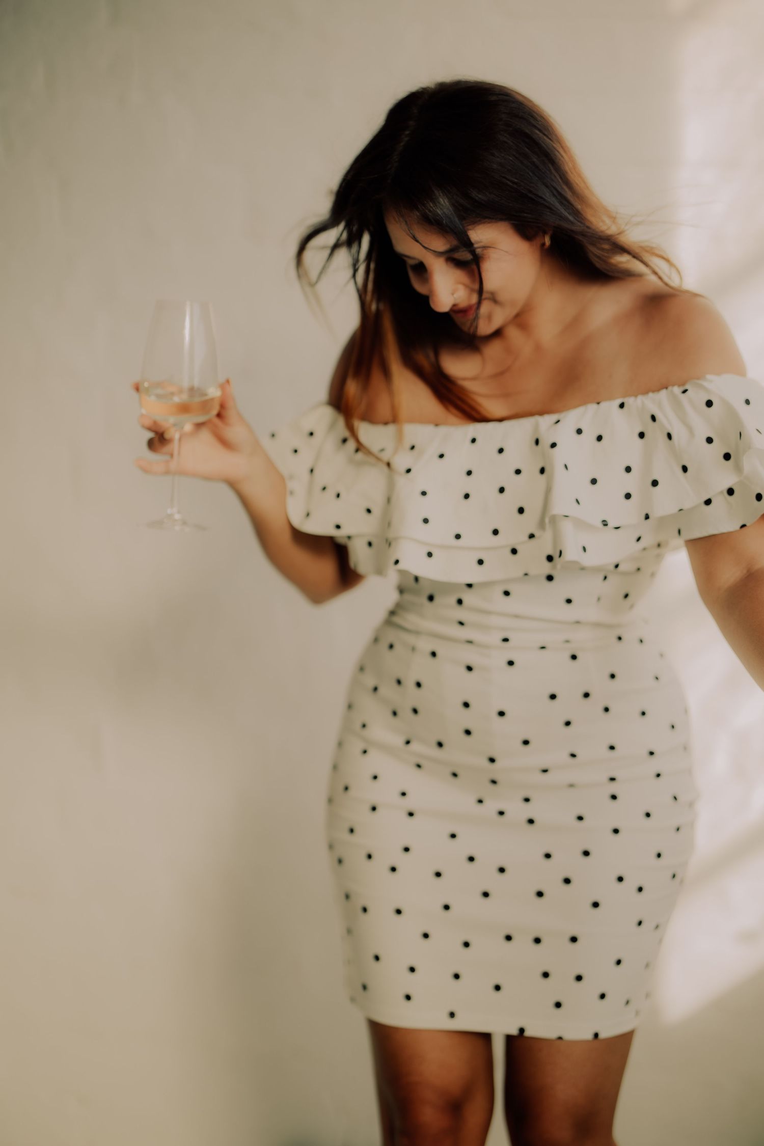 Woman in white polka dot off-shoulder mini dress holding a champagne glass, posing against a neutral wall.