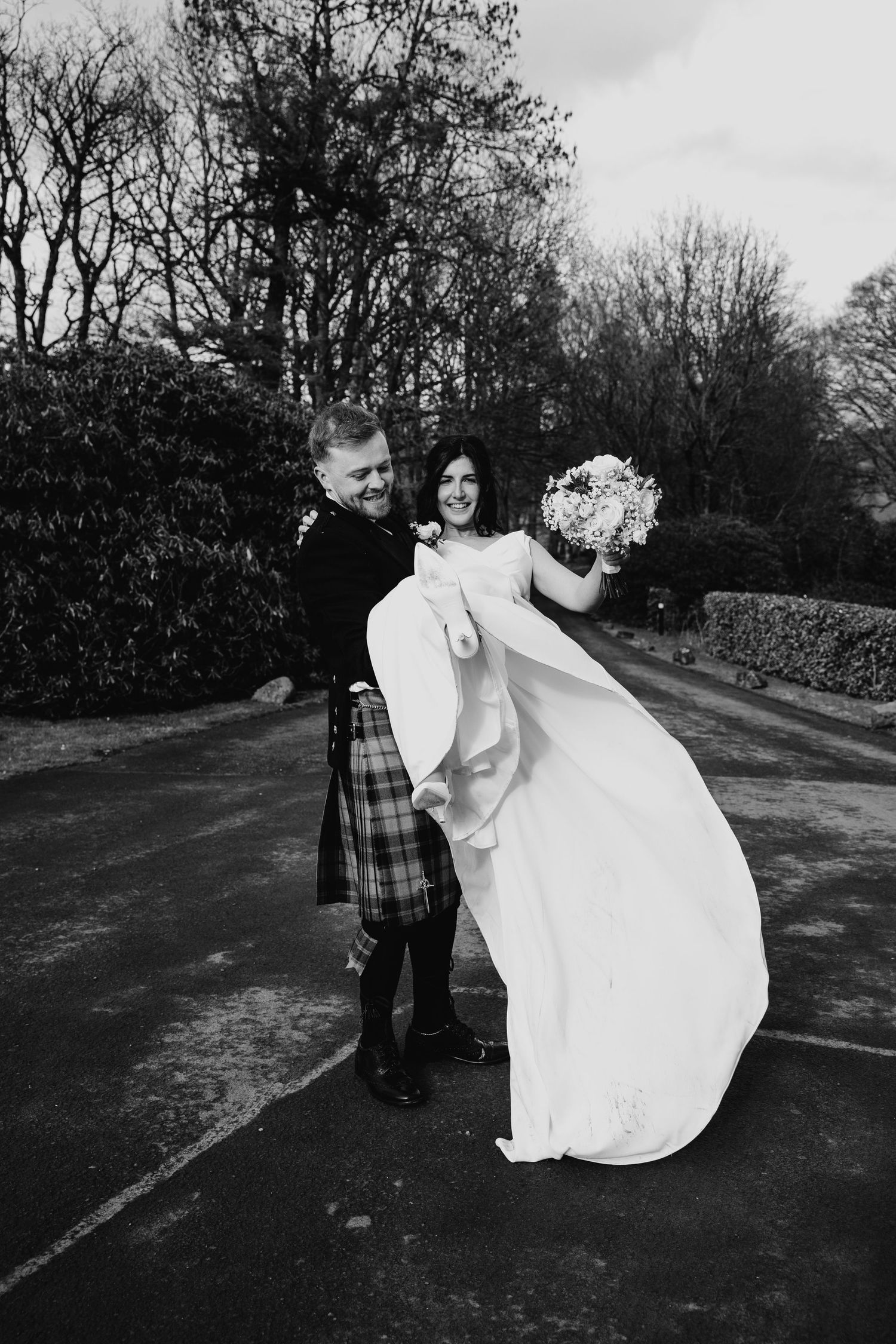 Black and white photo of groom in kilt lifting bride in flowing white gown outdoors at wedding.
