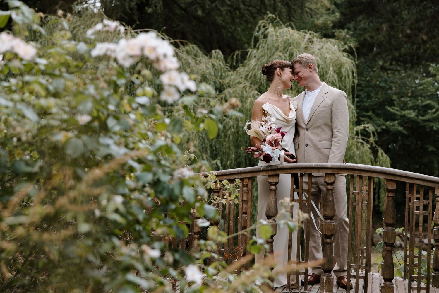 Newlyweds share tender moments on a wooden bridge surrounded by lush garden foliage.