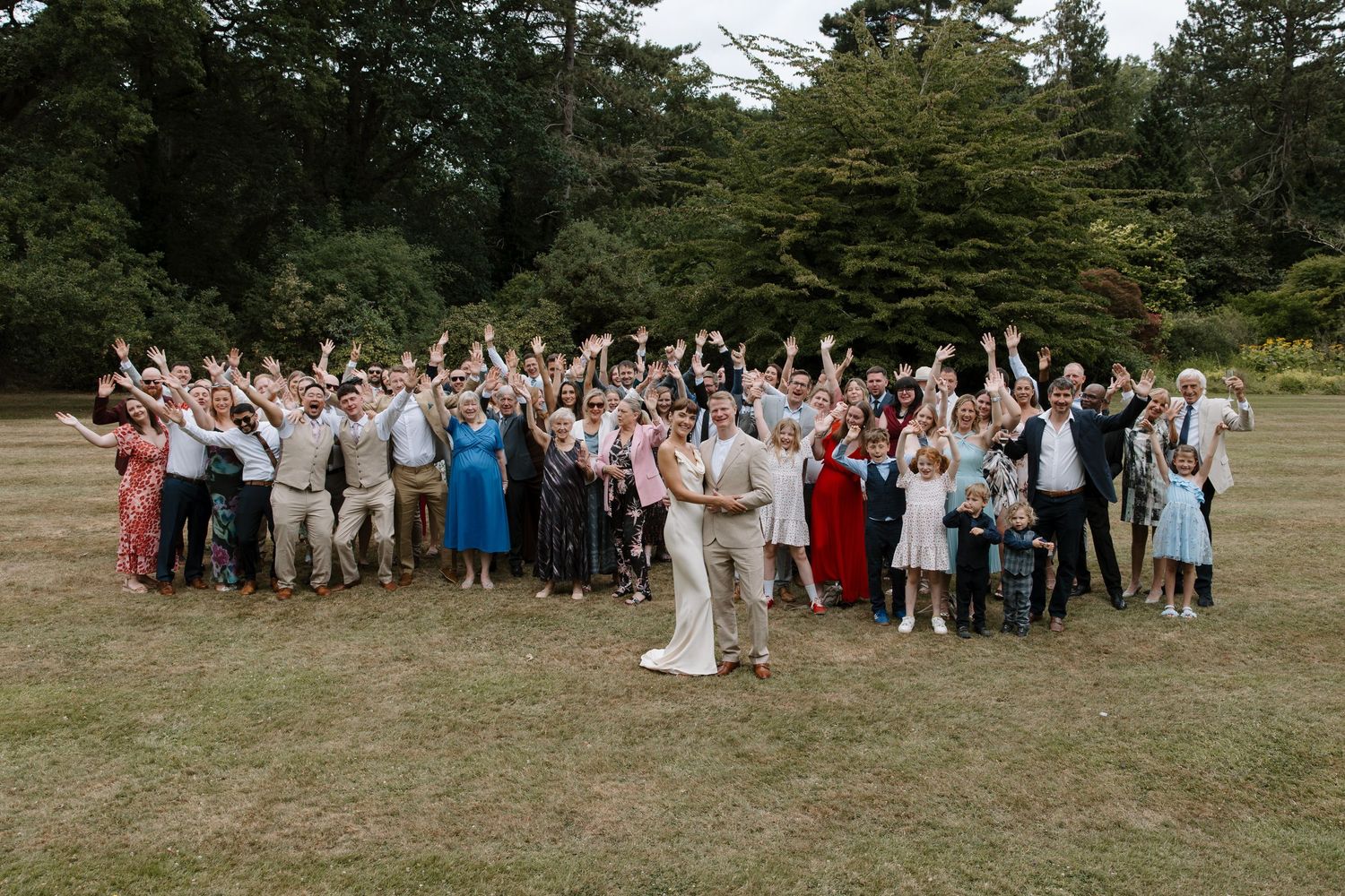 Large wedding group photo with guests gathered outdoors on grassy field.