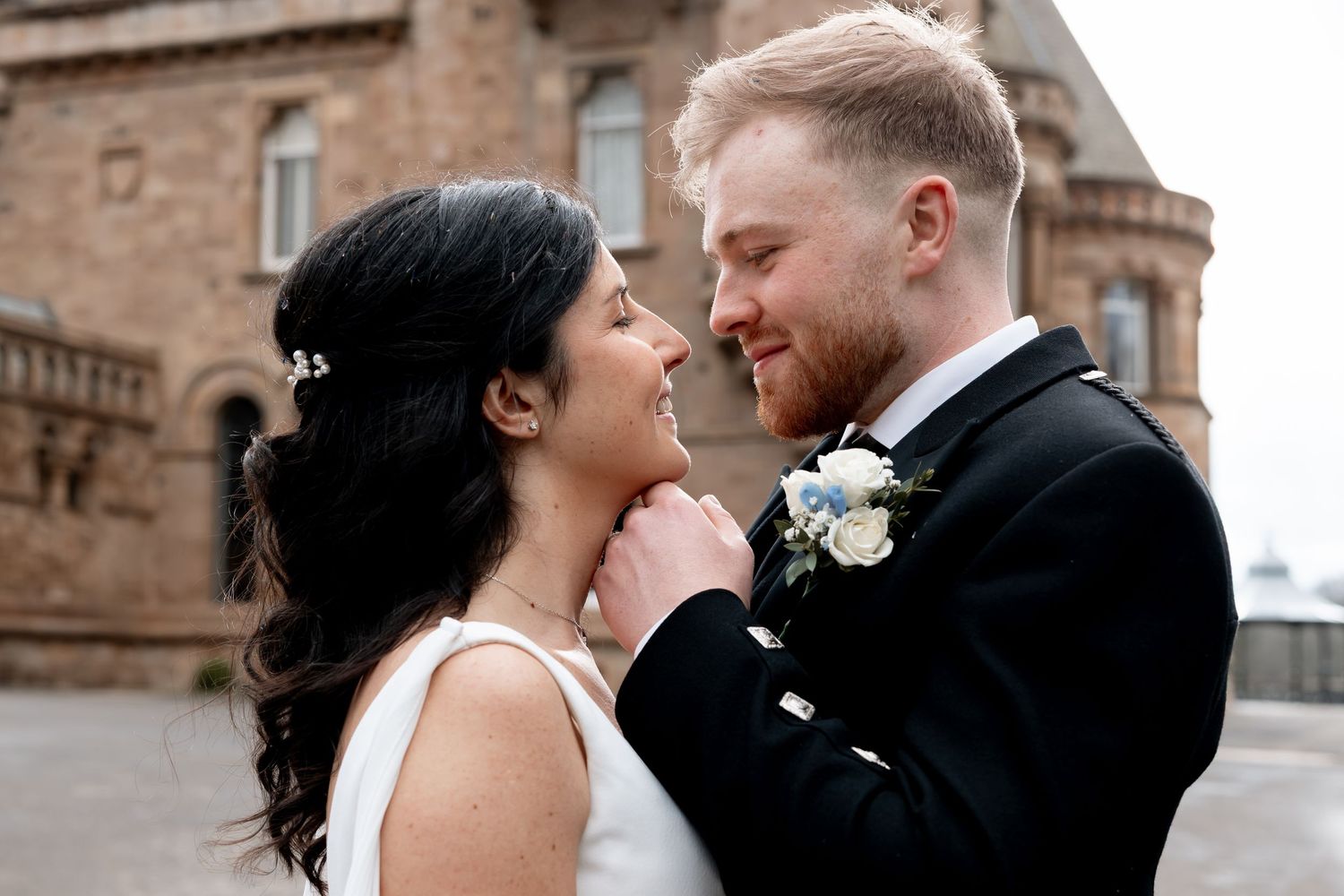 A bride and groom share a romantic moment, the groom tenderly holding her chin, both dressed in wedding attire outdoors.