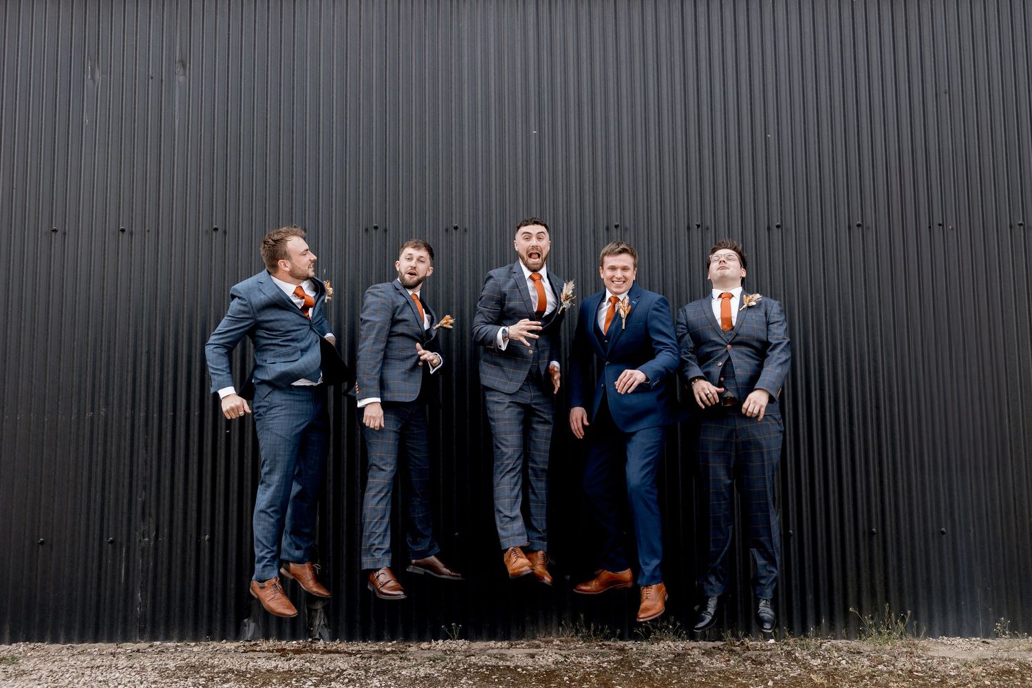 Groomsmen in matching suits pose playfully against dark wall showing off their brown shoes.