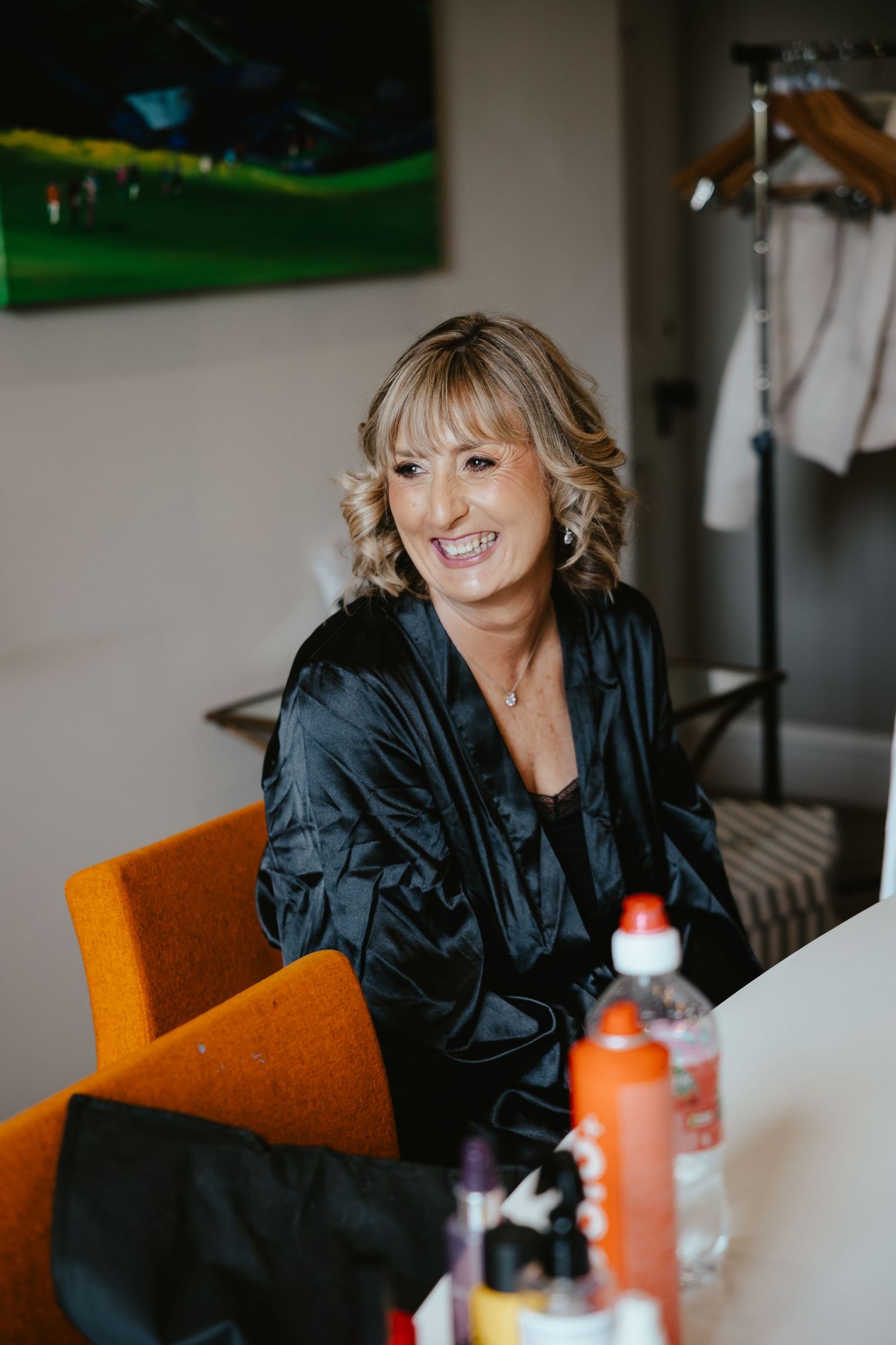 Smiling woman in black satin robe sitting at a table with beauty products, colorful art on wall behind her.
